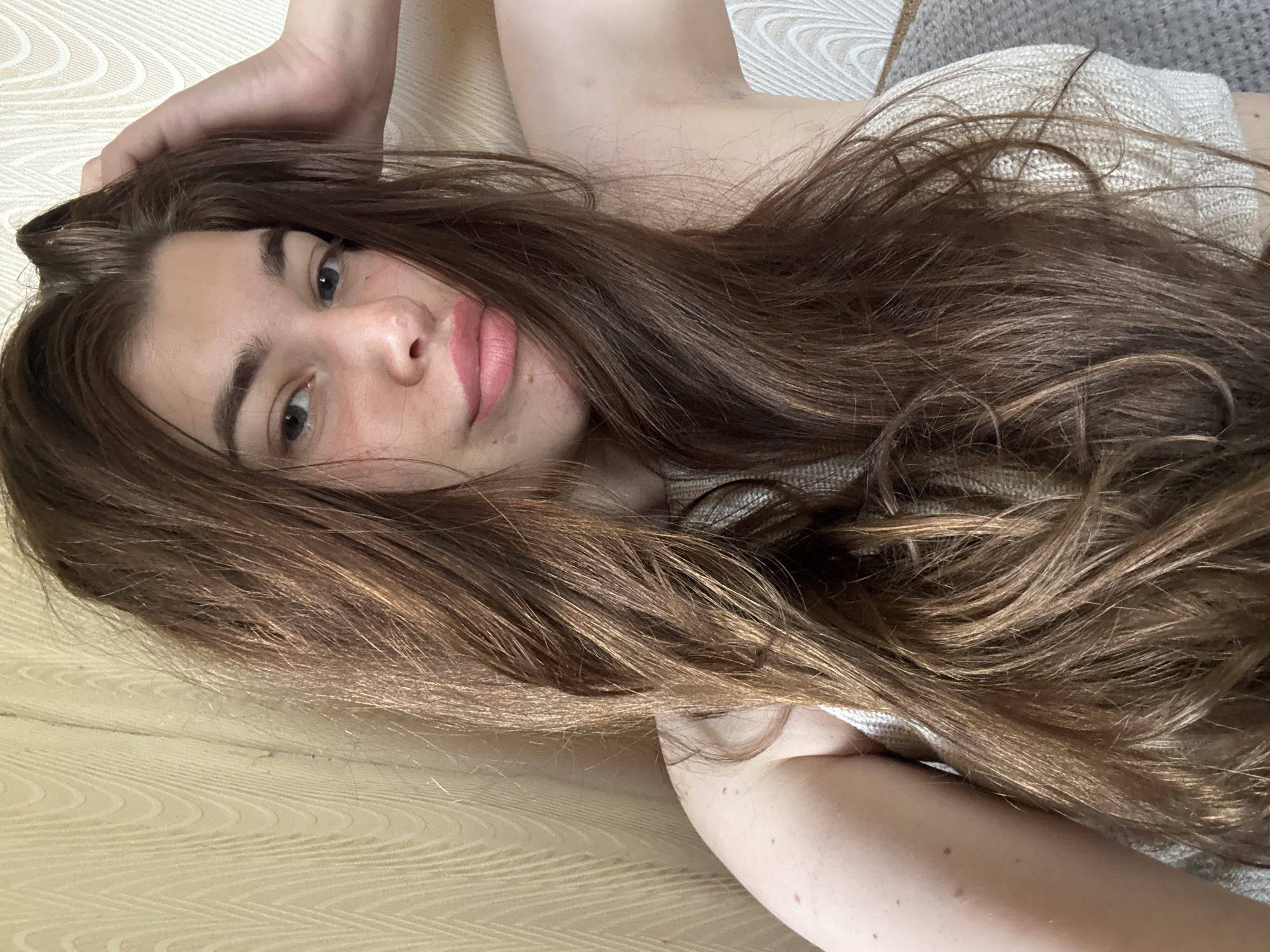 A young woman with long, brown hair is captured in a close-up, slightly angled selfie. She has light blue eyes and full, pink lips. Her right hand is raised and touching her head, near her hair. She is wearing a light-colored, textured top. The background consists of a wall with a subtle wavy pattern. The lighting appears to be natural and soft, suggesting an indoor setting. The image does not provide any specific clues about the location within Мещёры, Russia.