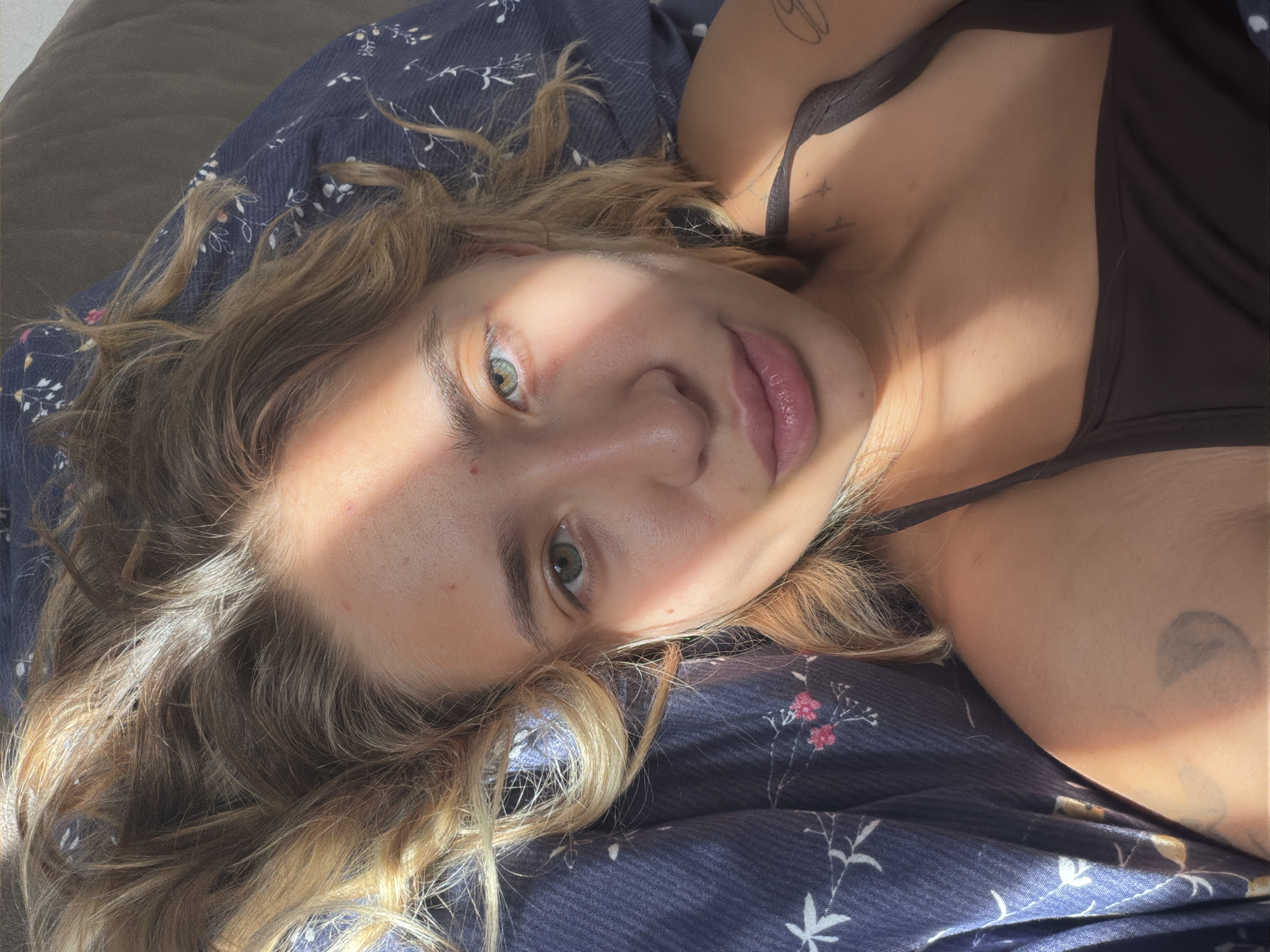 A young woman with blonde hair lies on her back on a blue floral patterned sheet. She is looking at the camera with her right eye clearly visible and her left eye partially obscured by a shadow. Her lips are slightly parted. A warm light illuminates her face from the left side. She is wearing a dark brown camisole. There are two small tattoos visible on her shoulder and upper chest area. The image is taken from a high angle.