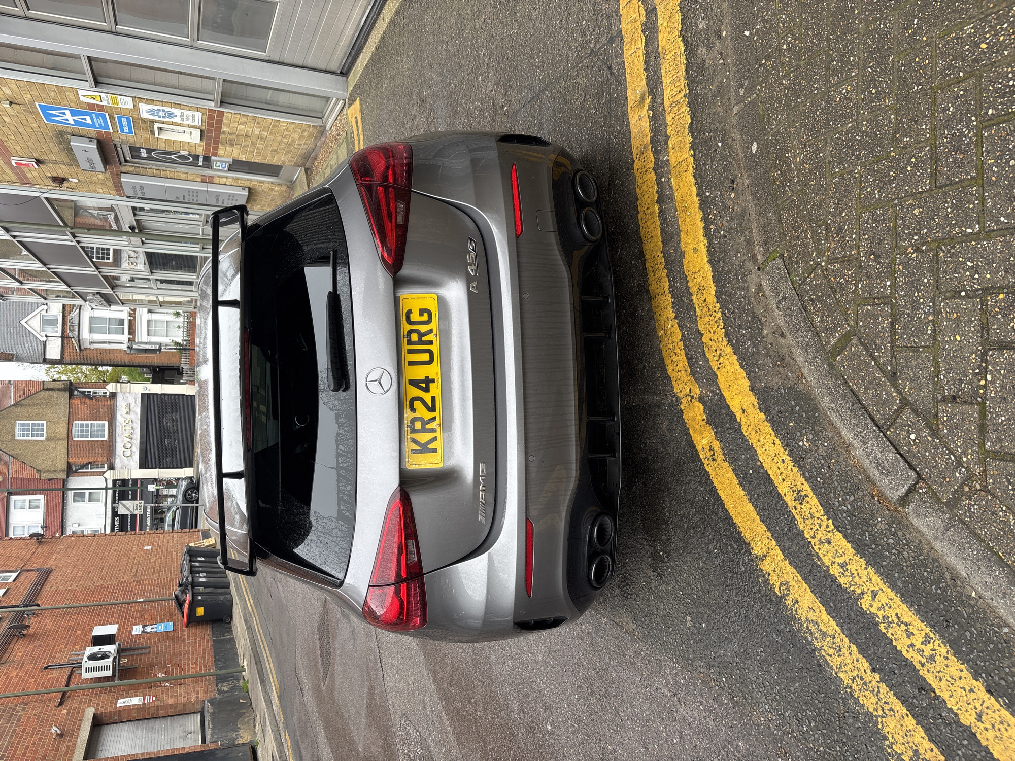 A silver Mercedes-AMG A45 S car, registration number KR24 URG, is parked on a street in an urban environment. The car is positioned alongside double yellow lines on a wet road surface. The surroundings include buildings made of brick and other materials, some with commercial signage. There are refuse bins, and what appears to be an air conditioning unit visible near the brick building. The location is most likely in London, United Kingdom based on the streetscape and the car's license plate.
