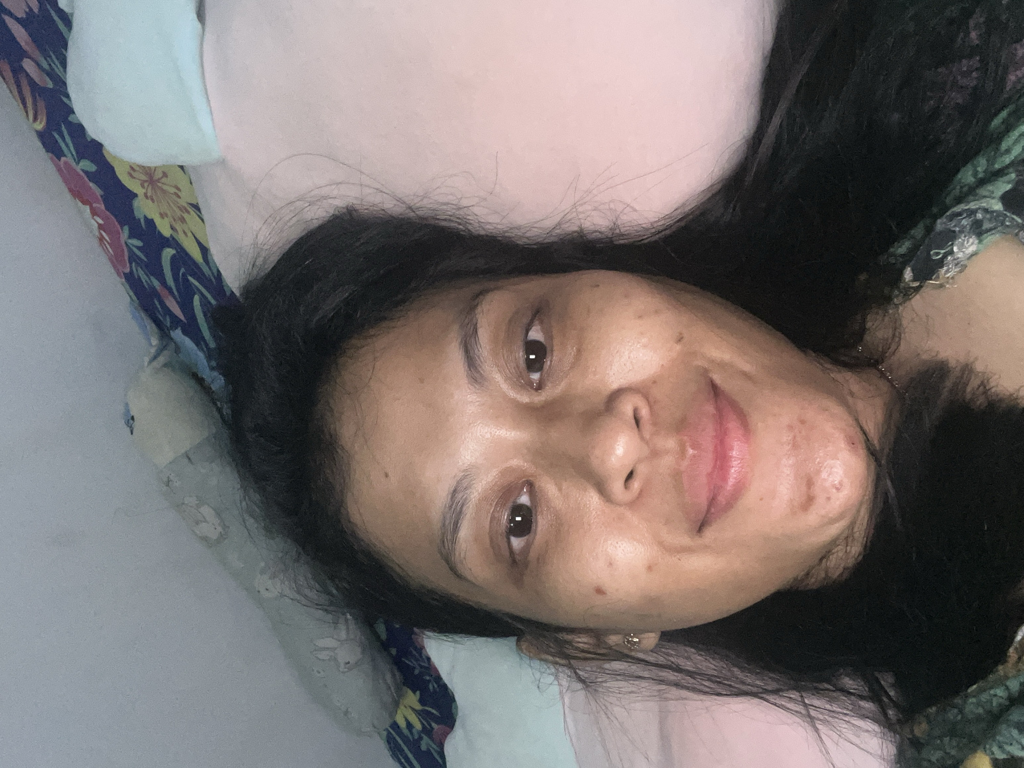 The image is a close-up, low-angle selfie of a woman lying down, with her head resting on pillows. She has dark hair, brown eyes, and her skin shows some texture and blemishes. She is wearing a dark green top with a floral pattern. The background consists of several pillows, some solid pastel colors and one with a floral pattern, and a bolster pillow with a light grey cover featuring cartoonish animal prints. The lighting appears to be artificial and dim. The scene is indoors, likely a bedroom, and there is no indication of the specific location within Kecamatan Pati, Indonesia, beyond it being an interior residential setting.