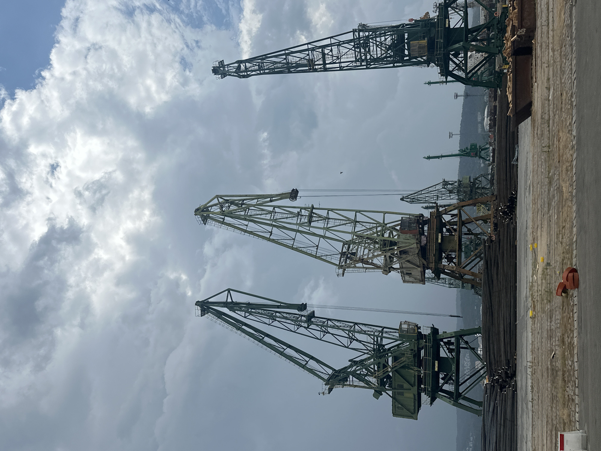 The image depicts a cloudy sky over a harbor with several large industrial cranes. In the foreground, a large pile of dark, cylindrical objects, likely rebar, is stacked on a concrete surface. Further back, more rebar is visible, along with several metal containers, some filled with what appears to be wood scraps. The cranes, which are green and metal, vary in size and design. The largest cranes are in the middle ground, with their booms extended. In the background, a hilly, urban landscape is visible under the overcast sky, with some buildings and more distant cranes. The overall scene suggests an active industrial port or shipyard.