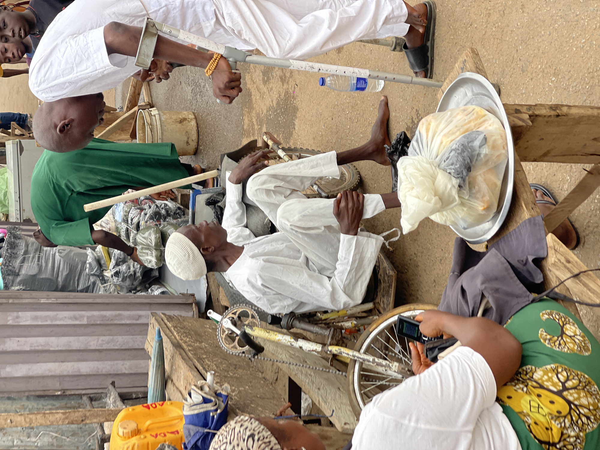 In Kuchingoro, Nigeria, the scene features an individual reclined amidst a bicycle frame and scattered materials, wearing a white outfit and a white skullcap. The individual appears to be interacting with others. One person stands nearby, utilizing crutches, while another, wearing a green shirt, is also present. There's a tray filled with bagged goods in close proximity. Surrounding the scene are various objects including a water bottle, assorted containers, and wooden planks. Another individual is using a cell phone, wearing a white shirt and green patterned fabric. The background and foreground consist of a mix of urban elements and signs of activity.