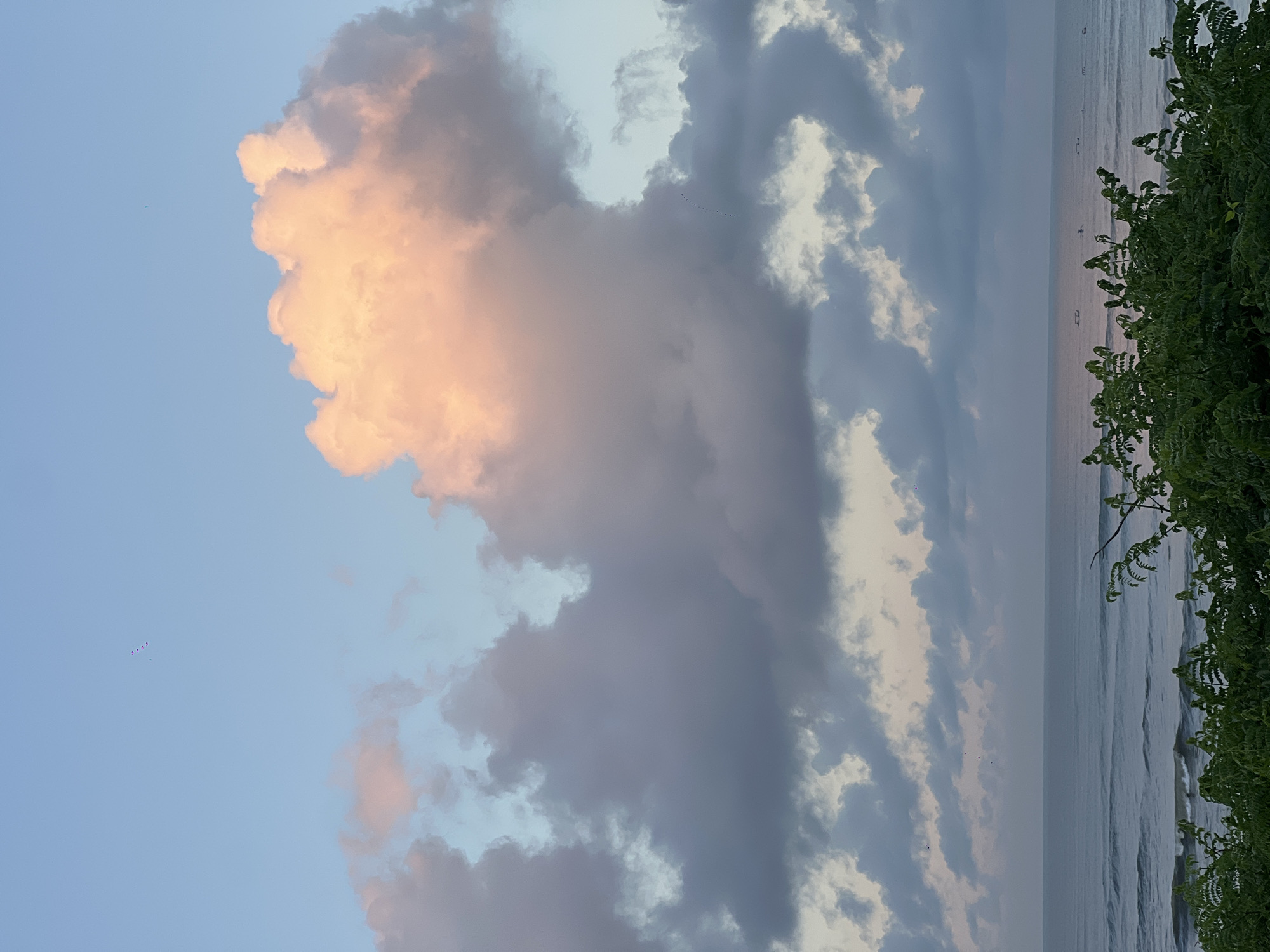 The image shows a coastal scene with a view of the ocean and sky. The sky is filled with clouds, some illuminated with a pinkish hue, possibly from the setting sun. The ocean appears calm with small waves. On the right side of the image, there is green foliage. Several boats on the water are visible. The location is Unknown, Indonesia.
