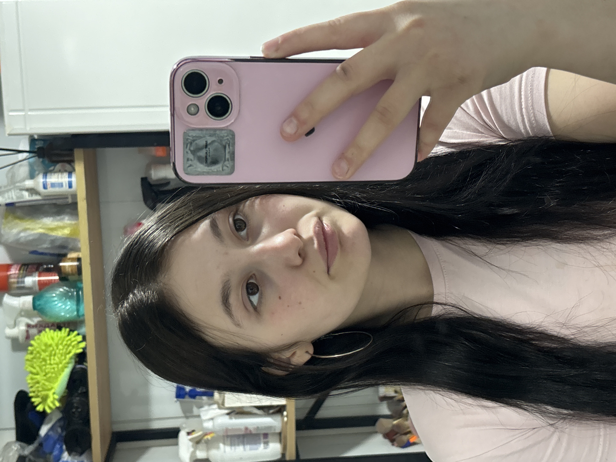 A young woman is taking a selfie with a pink iPhone. The phone is in her right hand, partially obscuring her face. She has dark hair and is wearing a light pink t-shirt and hoop earrings. The background shows a cluttered bathroom shelf with various cleaning products. The location is not discernable.