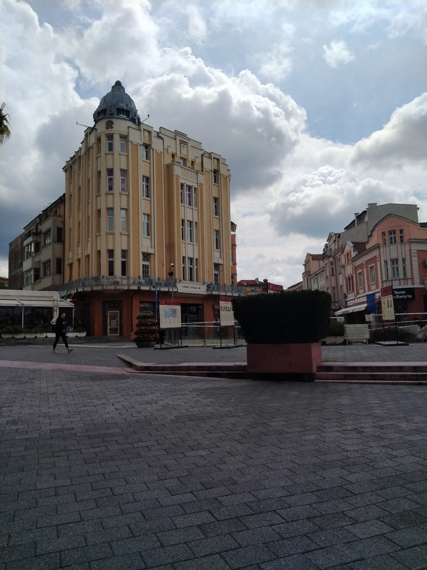 The image presents an urban scene in Plovdiv, Bulgaria, on an overcast but bright day. The main subject is a grand, multi-story building with a light yellow/beige facade, located on the left. This building features vertical architectural elements, numerous windows, and a distinctive dark dome capping its corner. A stylized "ST" logo is visible near the dome. On its ground floor, there's a cafe or establishment with outdoor seating. Above its entrance, the text "ОФИС СГРАДА ББ" (Office Building BB) is visible.
To the right, a street or pedestrian zone extends into the background, lined with an array of older buildings in varied styles and colors, including pink, white, and gray. One of these buildings prominently displays a sign reading "Tavex Валута" (Tavex Currency/Exchange).
In the foreground, the ground is paved with dark gray rectangular pavers. A large, neatly trimmed dark green bush in a reddish-brown planter box occupies the mid-ground. A single person, dressed in dark clothing, walks across the paved area from left to right. The sky is filled with large, fluffy white and gray cumulus clouds, though patches of brighter sky are also visible. Small dark specks, possibly birds, are scattered across the sky. The overall impression is that of an active city square or street.