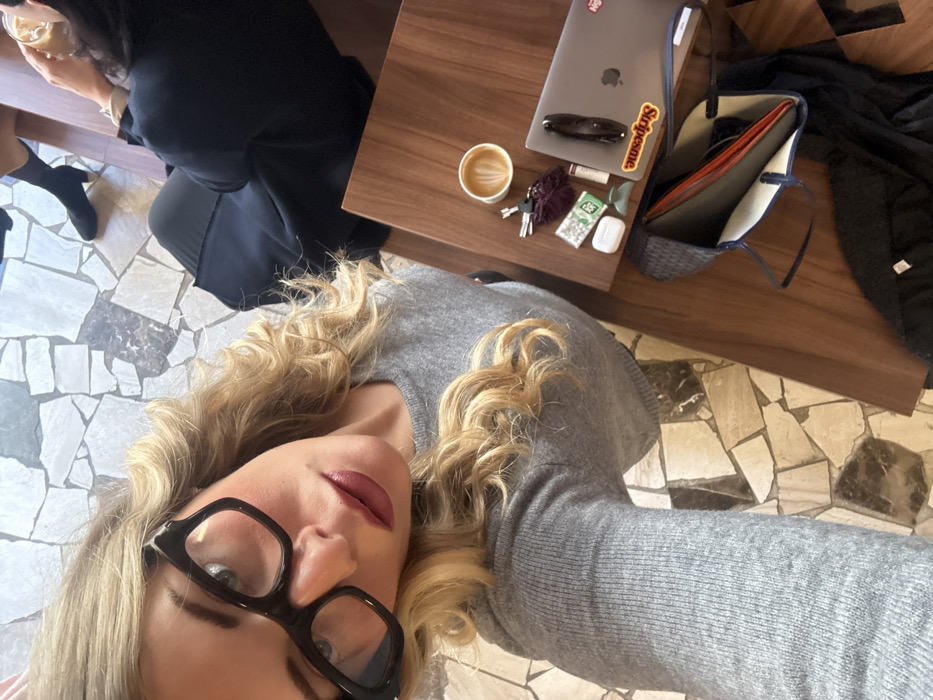 The image captures an indoor scene in the city of Paris, France, likely within a cafe or similar establishment, characterized by a mosaic tiled floor composed of irregularly shaped light and dark tiles. The primary subject, a person with blonde, wavy hair, wearing black-framed eyeglasses and a grey ribbed long-sleeved sweater, is visible from the chest up, looking directly into the camera. Dark lipstick is applied. To the right, a second individual, partially visible in dark clothing, holds a clear glass containing an iced beverage. A dark brown wooden table occupies the mid-ground, displaying a white paper cup of coffee with latte art, a set of keys with a dark purple fluffy keychain, a small white rectangular packet, white AirPods, a green hair clip, a silver laptop with a "Supreme" sticker and another red sticker, black sunglasses resting on the laptop, and a small cylindrical object. Below the table, a dark blue tote bag with an orange interior holds documents, and a dark jacket or garment is partially visible. Additional legs and feet in the background suggest the presence of other individuals.