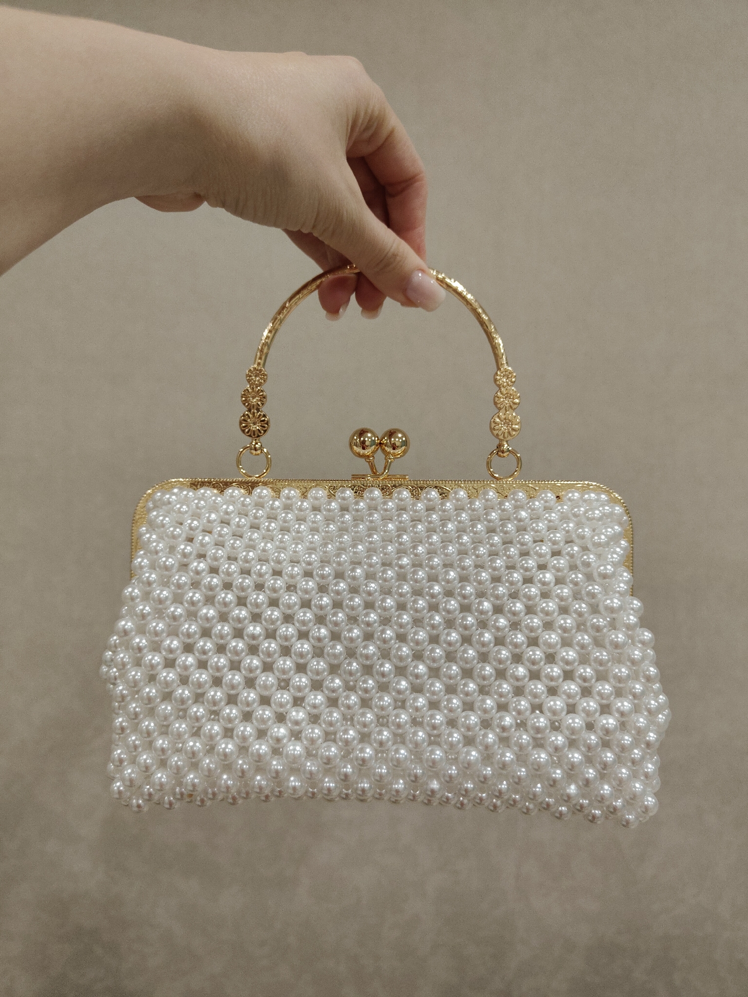 A hand is holding a white pearl-covered purse with a gold handle and frame. The background is a plain beige color. The location and city are unknown.