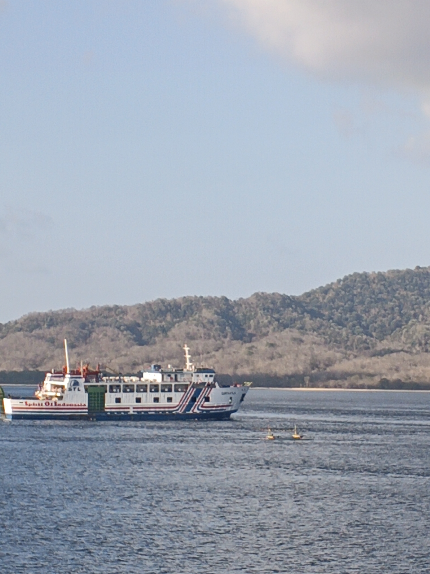 A ferry, labeled "Spirit Of Indonesia," travels across the water near the coast of Melaya, Indonesia. The ferry, white with red and blue accents, is large and appears to be a passenger or cargo vessel. In the distance, a densely wooded, mountainous island forms the backdrop under a hazy blue sky. Two small yellow buoys are visible in the water in the foreground, and the sea is a deep blue with visible ripples.