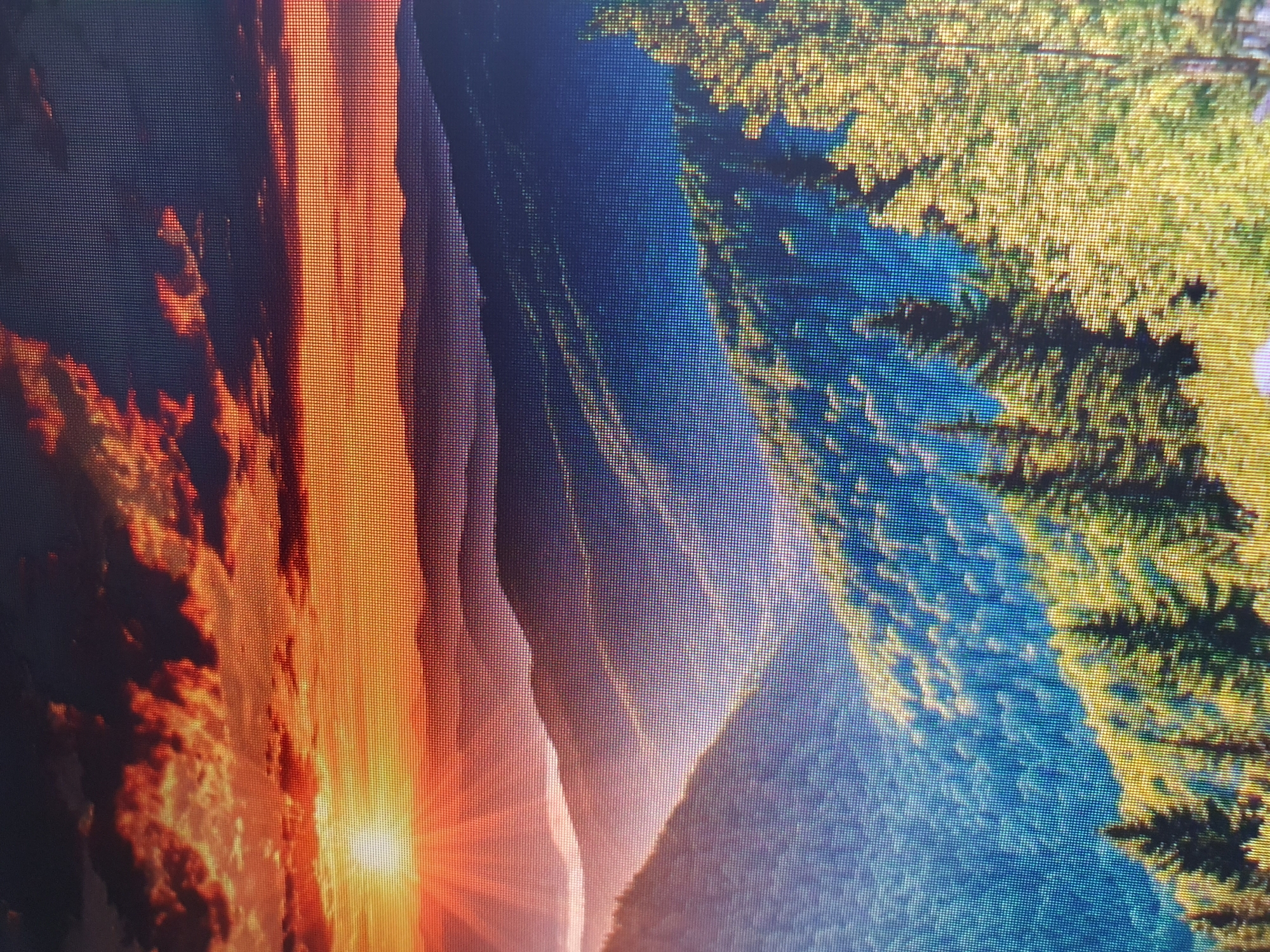 The image depicts a vibrant sunset over a mountainous landscape. The sky is ablaze with fiery oranges, reds, and yellows, with dark clouds scattered throughout. Sunbeams radiate outwards from the setting sun, illuminating the misty layers of mountains that recede into the distance. The foreground is dominated by lush green trees, some of which are evergreen pines, set against a backdrop of more distant, hazy mountain ranges. The overall impression is one of natural beauty and tranquility. There is no indication of the city of Tashkent, Uzbekistan, or any urban elements within the image.