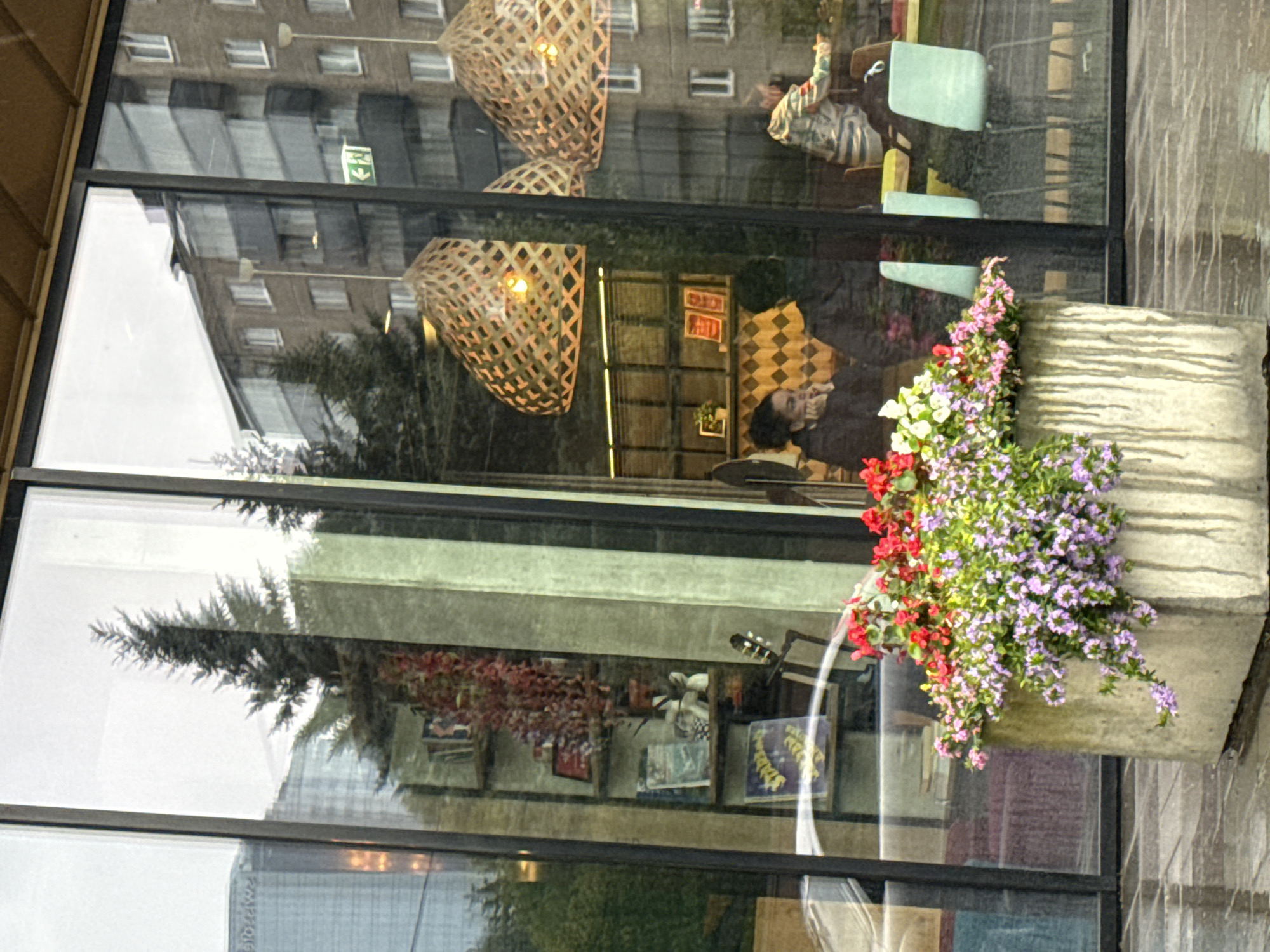 The image depicts a street-level view of a cafe or restaurant in Tallinn, Estonia, as evidenced by the distinctive architecture and the green exit sign visible through the glass, a common sight in European cities. The foreground is dominated by a large, concrete planter overflowing with a vibrant arrangement of pink and purple flowers, along with some red blossoms. The planter is positioned on a wet, reflective pavement, suggesting recent rain.
Through the large glass windows of the establishment, several elements are visible. On the left, reflections show trees and buildings, creating a layered visual effect. Inside, a shelf unit is stocked with books and decorative items, including a potted plant with cascading red leaves and what appears to be a small guitar or similar instrument.
Further inside, two individuals are seated at a table. One person, a woman with dark hair pulled back, is leaning on her hands, looking downwards. Another person, whose back is mostly to the viewer, is also seated at the table. In the background, another patron wearing a patterned jacket is visible, interacting with someone out of frame. The interior also features modern light fixtures with a woven design. The overall scene conveys a sense of urban life, with reflections and interior details blending to create a complex and somewhat blurred image.
