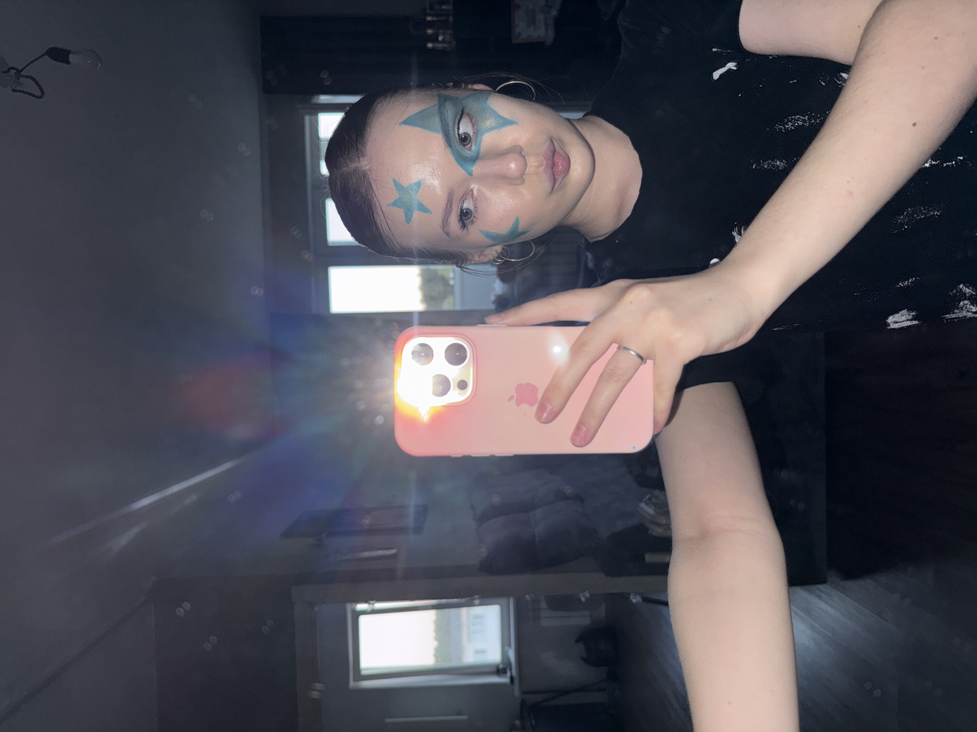 A young woman with face paint in the form of blue stars is taking a selfie in what appears to be an indoor setting. She is holding a pink iPhone in her right hand, its screen reflecting a bright light. She wears hoop earrings and a black t-shirt. The background is partially visible and includes a window with daylight streaming through, suggesting it's daytime. There is dark furniture or cabinets beneath the window, reflecting the light. The environment is a dimly lit interior with reflections, implying a polished surface. It's not possible to confirm the location in Chaikovsky, Russia.