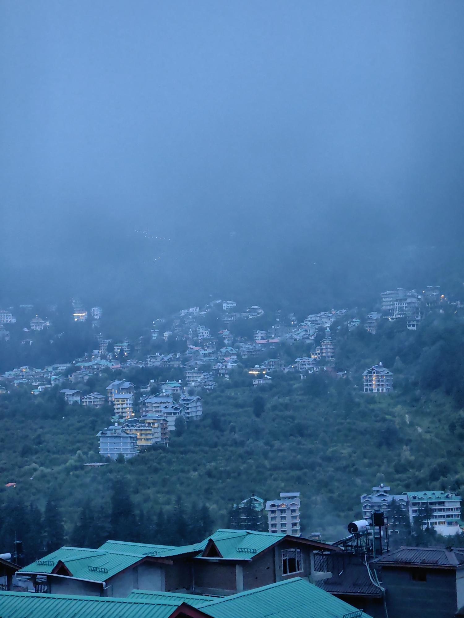 The image captures a high-angle view of a mountainous town, likely Vashisht, India, shrouded in a dense, misty atmosphere during twilight. The cityscape is built into the steep, forested slopes, characterized by multi-story buildings with predominantly light-colored facades and green-pitched roofs. Some buildings display illuminated windows, indicating occupancy. The terrain is rugged, with dense green foliage covering the hillsides between the structures. In the foreground, closer buildings with distinct green metal roofs are visible, some showing architectural details like dormer windows and visible plumbing fixtures on one rooftop. The overall mood is serene and slightly melancholic, with the mist softening the outlines of the buildings and creating a diffused light. There are no people or specific actions depicted in the scene.