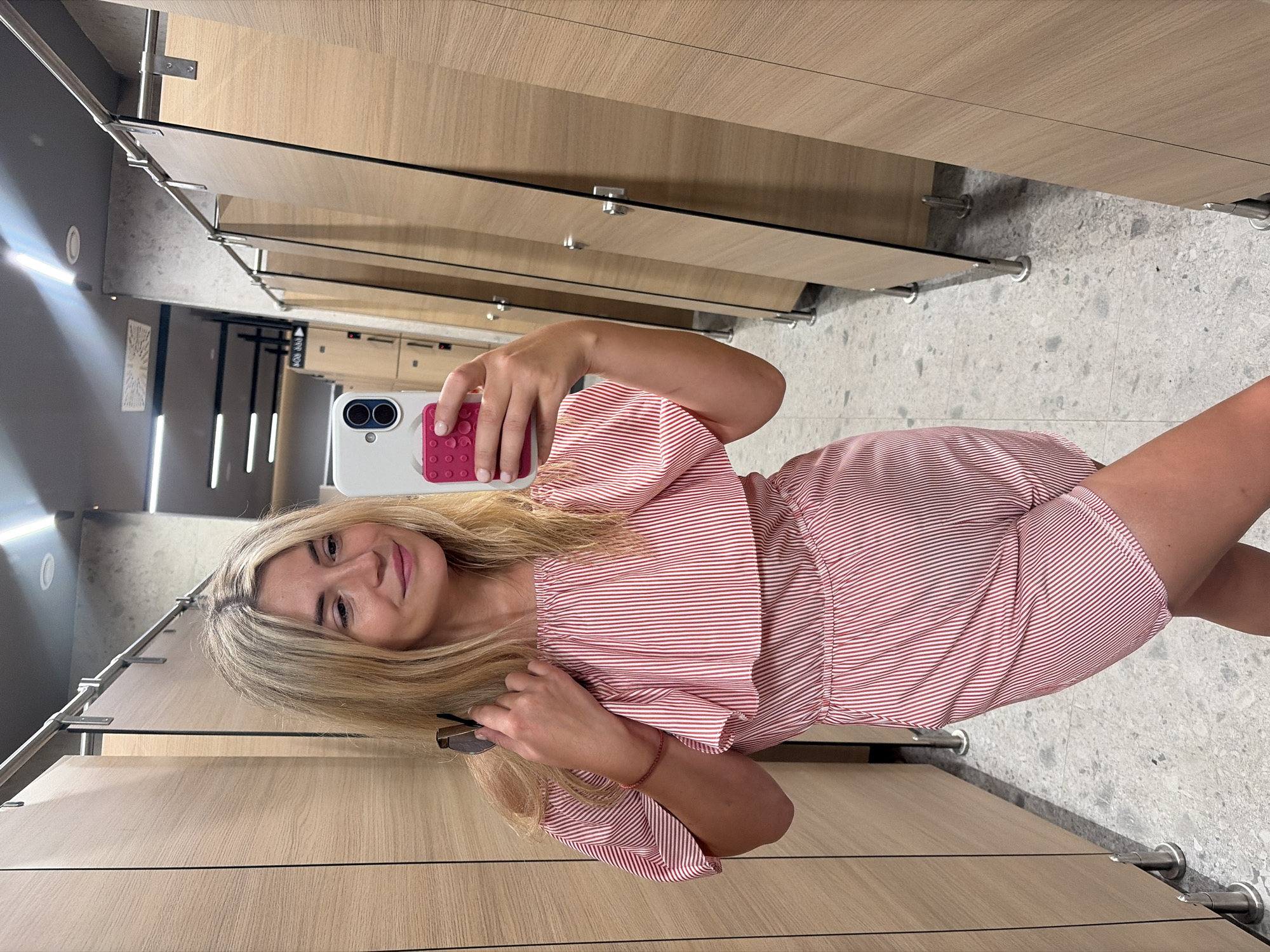 A blonde woman with fair skin and light brown eyes is taking a mirror selfie in what appears to be a fitting room or locker room. She is wearing a red and white striped off-the-shoulder romper and holding a smartphone in her right hand, which has a pink grip case. Her left hand is holding a pair of sunglasses and she is wearing a thin red bracelet on her left wrist. The room has a modern aesthetic with light wood paneled walls, chrome accents, and grey speckled flooring. The lighting is bright, with fluorescent lights visible in the ceiling. The background shows a row of similar fitting rooms, suggesting a commercial establishment. The location is identified as Podgorica, Montenegro.