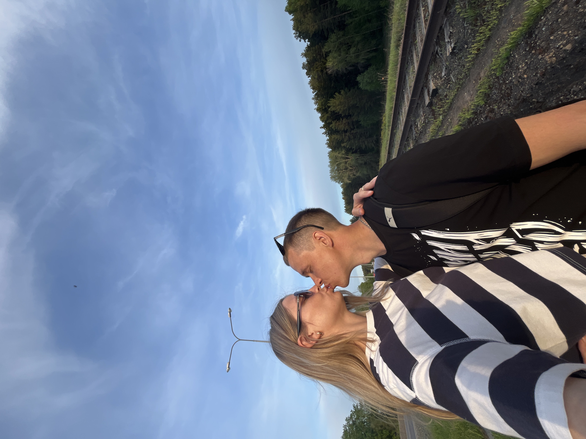 Two people are kissing. The person on the left has blonde hair and is wearing a striped shirt and eyeglasses. The person on the right has short dark hair and is wearing a black t-shirt and a chain. Sunglasses are perched on the person on the right's head. They are standing next to train tracks and the photo is being taken from below, looking upwards towards the sky. A lamppost is visible in the bottom left. The photo was taken in Chaikovsky, Russia.