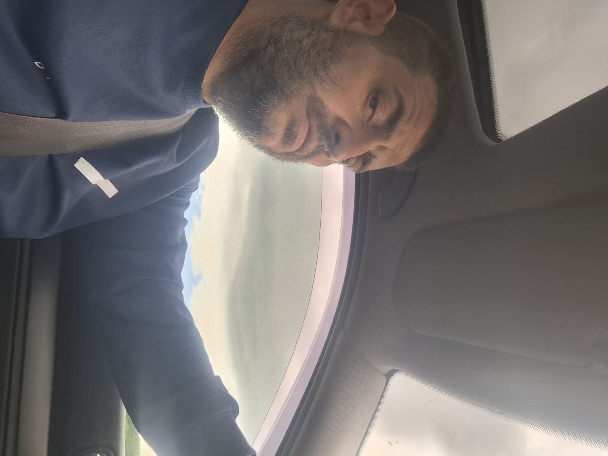 A man with a beard is seen from the chest up, inside a car. He is wearing a dark blue sweatshirt with a white rectangular patch on the left breast. A grey seatbelt is visible across his chest. The man is looking towards the camera with a serious expression. The interior of the car, including the sunroof and window frame, is visible. The outdoor environment appears cloudy with some hints of blue sky. The image was taken in Dolno Sahrane, Bulgaria.
