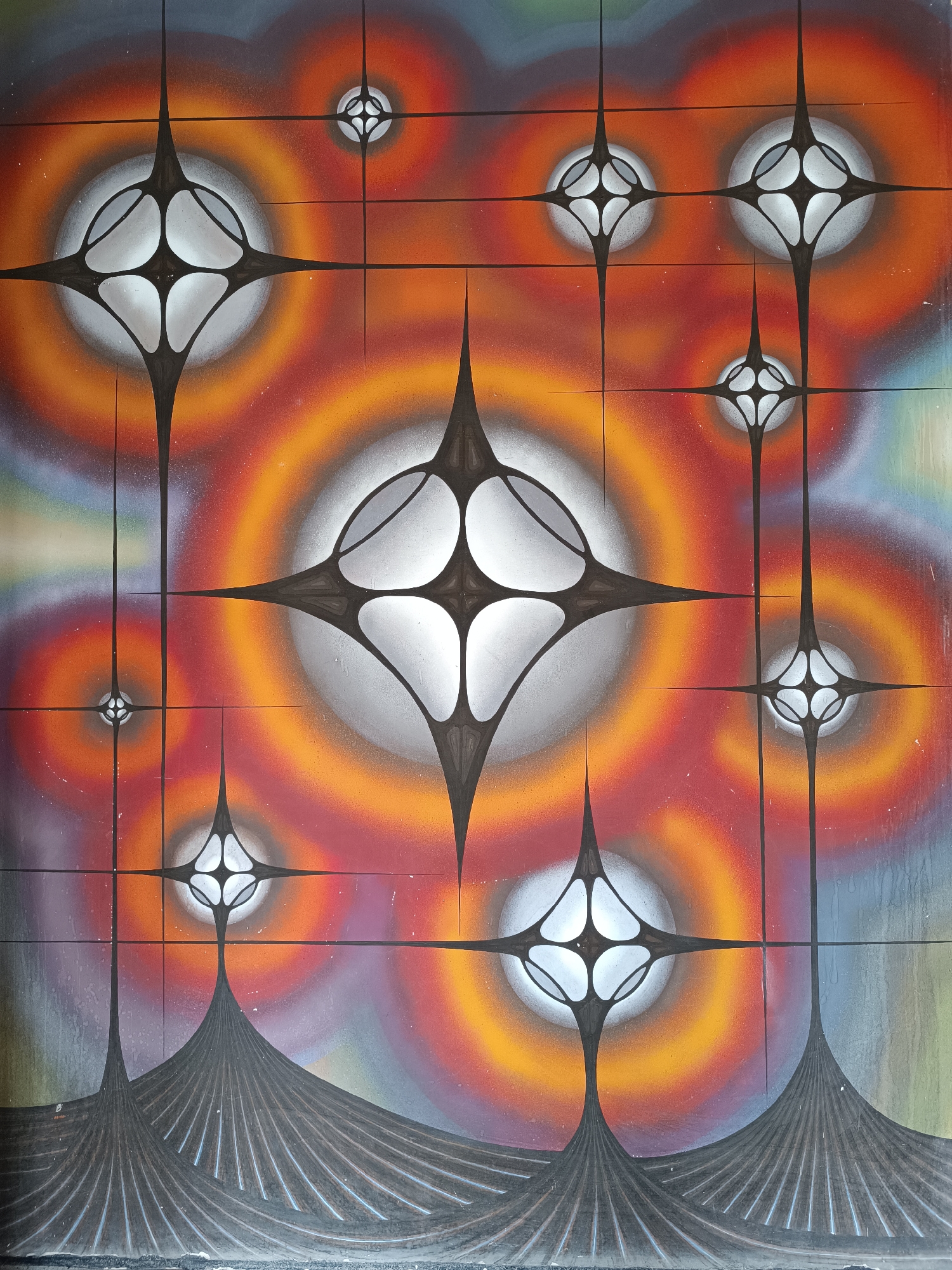 The image is an abstract painting or mural, possibly located in Sofia, Bulgaria (based on image context). It features a grid-like structure imposed over a background of concentric circles in varying shades of red, orange, yellow, green, and blue. Within the grid, there are several stylized star-like shapes with rounded edges, colored in shades of gray and white, with black outlines. These shapes vary in size, with the largest one near the center. At the bottom of the painting, there are stylized, vertically striated, dark gray shapes that resemble mountain peaks or abstract landforms. The painting appears to be on a textured surface, possibly a wall or canvas.