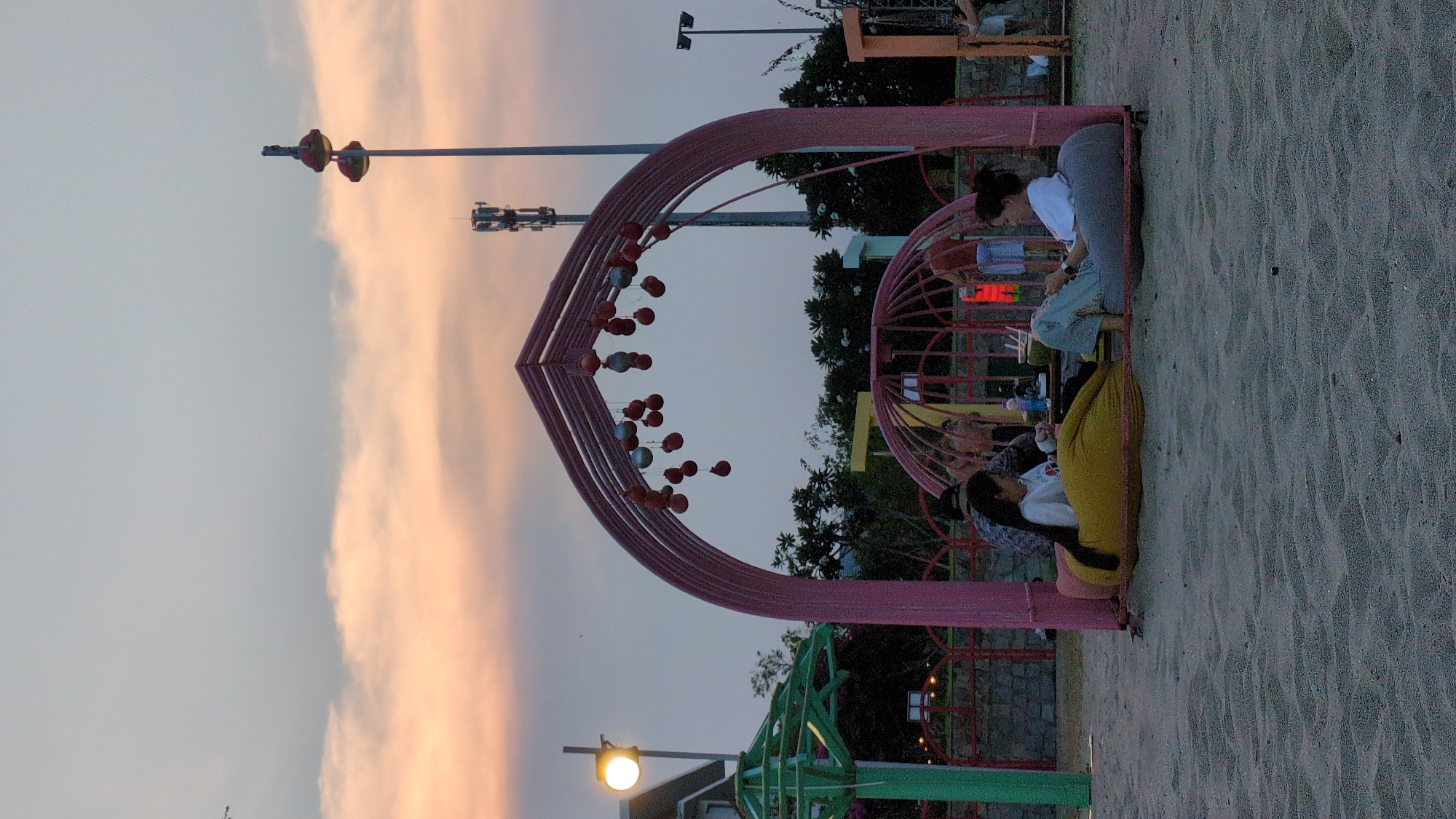 A nighttime outdoor scene at a playground or beach in Khánh Hải, Vietnam. Two women and a child are seated within a pink metal archway decorated with hanging spheres, possibly for a photo opportunity. One woman is kneeling, the other is sitting, and the child is lying down on a yellow beanbag. There are trees, lighting fixtures, and telecommunication towers in the background. The ground is sandy. The sky is overcast with a pinkish hue on the horizon.
