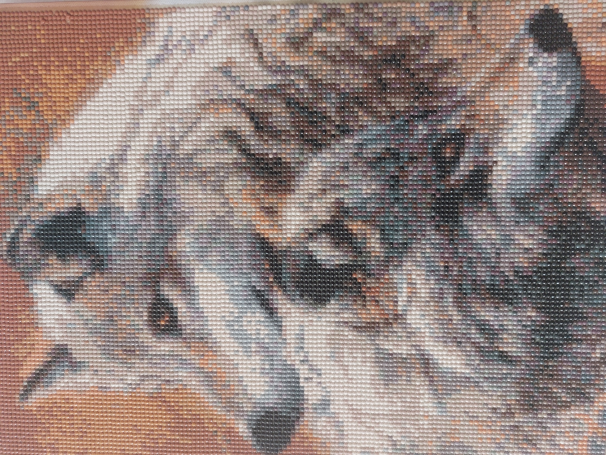The image displays a close-up view of a diamond painting, likely a wolf's head and upper body, rendered in a mosaic of small, colored resin beads. The painting is characterized by its pixelated appearance, with individual beads forming the intricate details of the wolf's fur, eyes, and snout. The color palette ranges from warm earthy tones for the background to cooler grays, whites, and blacks for the wolf itself, with subtle hints of purple and blue in the shadows. The texture of the beads is evident, creating a shimmering, faceted surface. The image focuses on the artistic representation of the animal, with no discernible urban or environmental context. The painting does not depict any specific location within the city of Sad, Ukraine, as it is a standalone artwork.