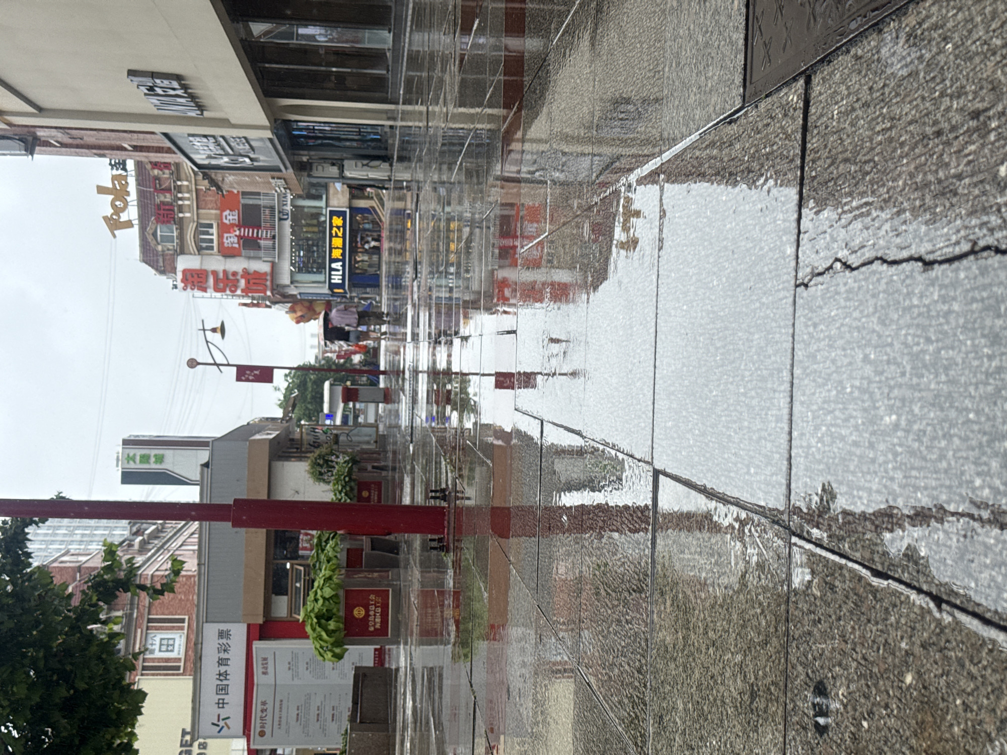 The image shows a wet, tiled pedestrian walkway in Qinhuangdao, China, on a cloudy day. The tiles are wet and reflective, showing the reflections of buildings and streetlights. In the background, there are several buildings with various signs in Chinese. One sign clearly reads "中国体育彩票" which translates to "China Sports Lottery". Another sign on a building in the distance says "HLA 海澜之家" which translates to "HLA - Home of the Sea". Two people are walking away from the camera, one holding an umbrella. A red pole is in the foreground, with plants in pots on either side. The sky is overcast, suggesting it has recently rained or is about to rain.