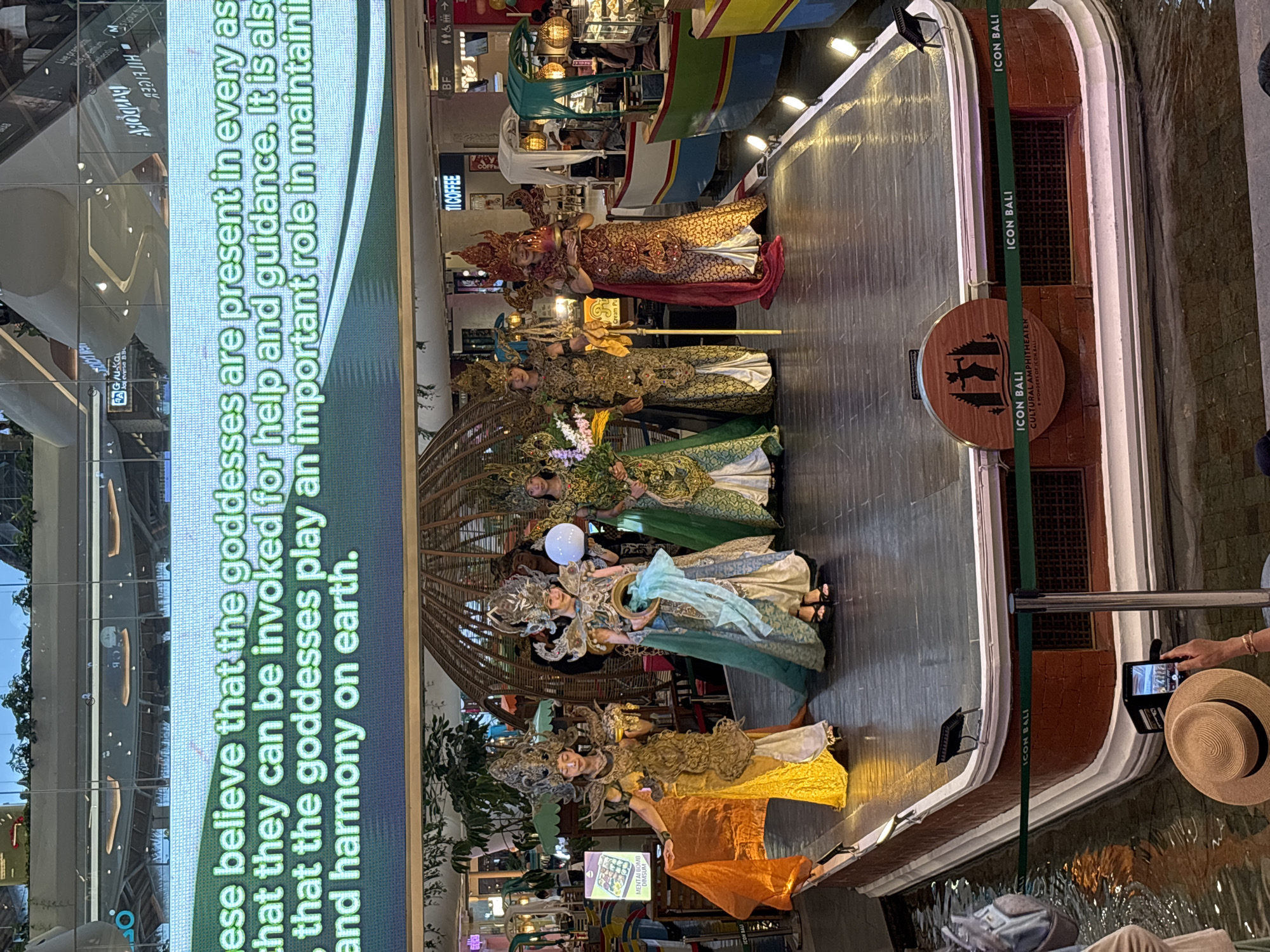 The image depicts a cultural performance at Icon Bali, a location in Denpasar, Indonesia. A group of individuals dressed in elaborate, traditional costumes, likely representing goddesses, are posing on a raised platform that resembles a boat. The costumes are ornate, featuring headdresses, intricate patterns, and flowing fabrics in vibrant colors like gold, orange, and blue. Some individuals hold props such as orbs, a golden ring, or flowers. Behind the performers, a large digital screen displays text in green against a dark background, with some of the text legible, reading, "...believe that the goddesses are present in every as... that they can be invoked for help and guidance. It is also... that the goddesses play an important role in maintainin... and harmony on earth." The platform is situated over water, with decorative lighting illuminating the scene. In the background, parts of a shopping mall environment are visible, including shops and people. A person in the foreground is taking a picture of the performance with a smartphone. The overall atmosphere suggests a cultural or promotional event aimed at showcasing Balinese traditions.