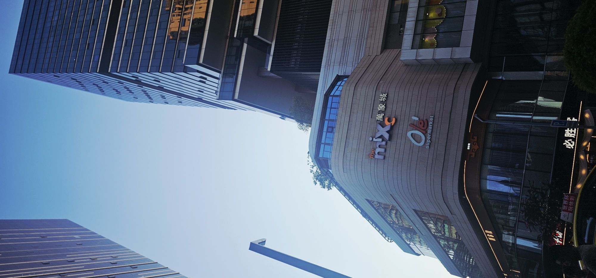 The image shows a low-angle view of modern urban architecture in Jiulongpo District, China. The sky is clear and bright blue. On the right, a tall, modern skyscraper with dark glass windows and horizontal detailing dominates the frame. To the left and slightly in the foreground, a curved building with a textured, light-colored facade is visible. This building features prominent signage: "the mixC" in colorful lettering, followed by Chinese characters, and below that, "Ole' supermarket" in white and orange. The "Ole' supermarket" sign is illuminated. The facade of the curved building has large windows reflecting the sky and adjacent structures. A street lamp with a distinct geometric shape is positioned in the middle left of the image, pointing upwards. At the bottom of the curved building, storefronts are visible, some with illuminated signs in Chinese characters, and one appears to be a parking sign. A yellow vehicle is partially visible in the lower left corner. Trees and greenery are present on and around the curved building, particularly on its rooftop and a lower level. The overall scene depicts a vibrant commercial district with contemporary architectural styles.