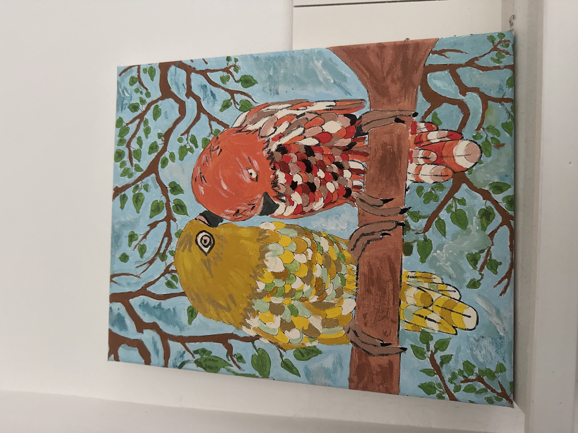 An eye-level, close-up shot captures a painting of two stylized birds perched on a brown tree trunk against a pale blue background. The tree branches extend outwards and are adorned with green leaves. The top bird is primarily red with white, brown, and black markings on its feathers. The bottom bird is yellow with white, yellow and green markings on its feathers. The painting is hanging on a white wall.
The information about the location in Plovdiv, Bulgaria is not visible in this image.