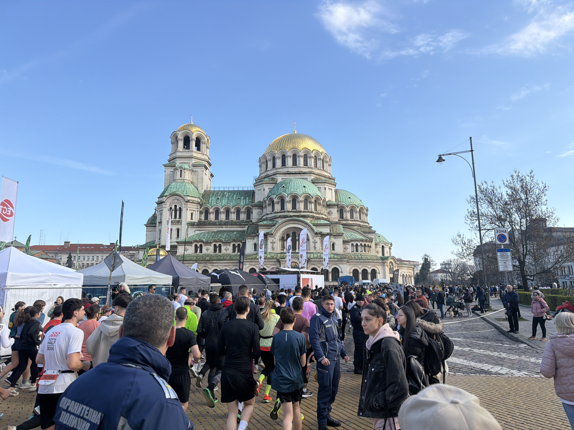 The image captures a crowded public space in Sofia, Bulgaria, in front of the Alexander Nevsky Cathedral, characterized by its gold domes and green roofs. A large group of people is present, many of whom are participating in a running event as indicated by their athletic attire and bib numbers. Some individuals appear to be spectators or event staff. Tents and banners are erected, likely related to the running event. The sky is clear and blue. The environment includes surrounding buildings, trees, a lamppost, and street signage.