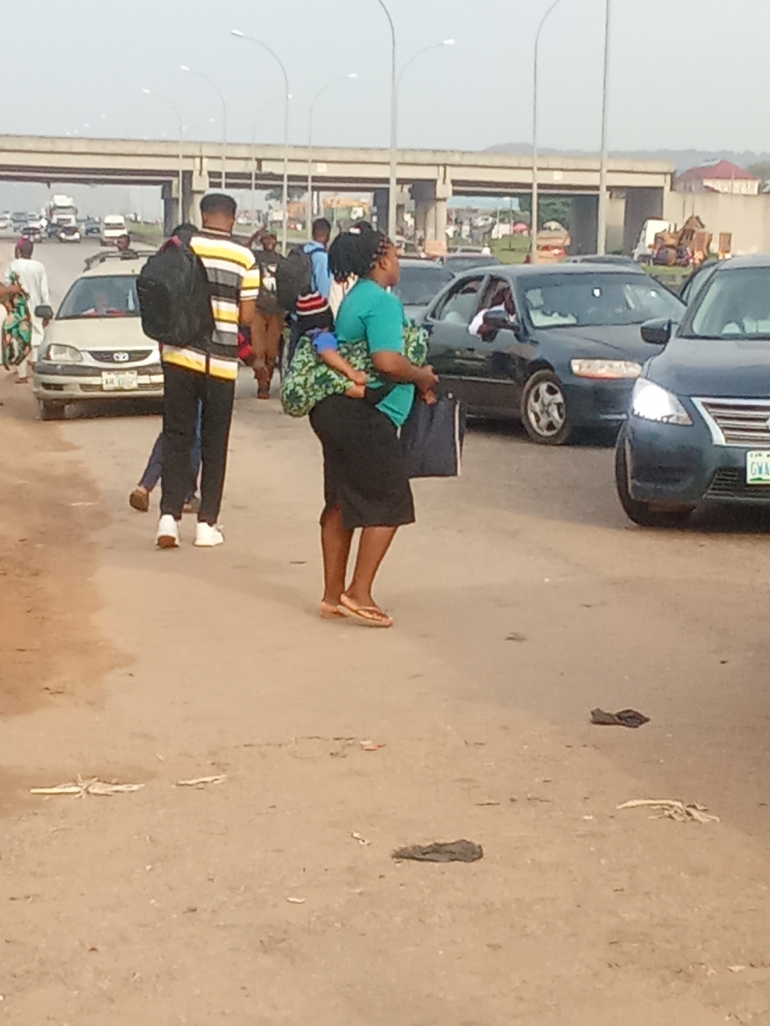 In Kagini, Nigeria, a woman wearing a turquoise top and a dark skirt, with her hair in braids, is seen from the chest down. She is carrying a baby on her back wrapped in a green and white patterned cloth and holding a dark blue bag in her left hand. She is walking on a sandy, dusty road with visible debris, suggesting an urban or semi-urban environment. Several vehicles are present, including a dark sedan in the foreground with its headlights on, and other cars and trucks in the background, indicating a road with traffic. A man with a backpack, wearing a striped t-shirt and black pants, is walking behind the woman and the baby, along with other pedestrians, some of whom are also carrying bags or backpacks. An overpass with streetlights is visible in the background, suggesting the scene is near a highway or major road intersection.