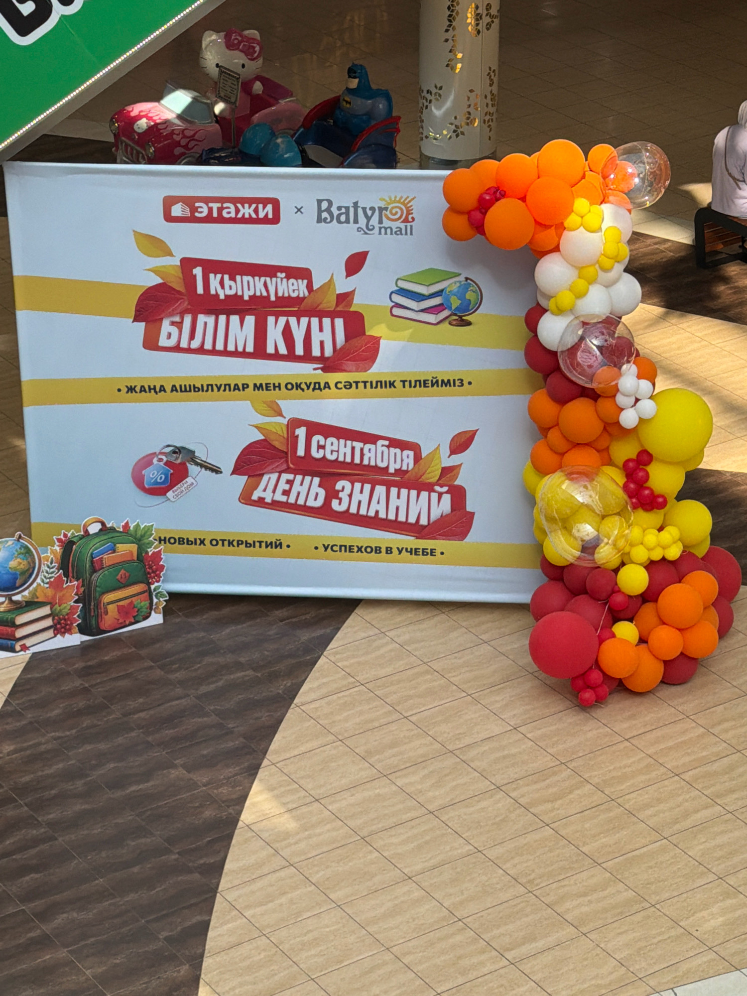 The image depicts a storefront or event display with a large banner and balloon decorations. The banner features text in Russian and Kazakh, indicating a sale or event related to the start of the school year. The Russian text "1 сентября День Знаний" translates to "September 1st, Knowledge Day," a common holiday in post-Soviet countries marking the start of the academic year. The Kazakh text "1 қыркүйек Білім күні" also translates to "September 1st, Day of Education."
The banner has logos for "Этажи" and "Batyrmall," suggesting this is a promotion within a shopping mall or store named Batyrmall. The visual elements include autumn leaves, books, a globe, and a backpack, all symbolic of education and the new school year. Balloon arch in vibrant colors like red, orange, and yellow is prominently featured, extending from the right side of the banner upwards.
In the background, there are decorative elements that appear to be part of the mall's interior, including a tall cylindrical pillar with an intricate pattern. To the far left, a green banner with white text is partially visible. In the upper left corner, there are toys including a Hello Kitty figure, a red car, and a Batman figure. In the far right, a person is seated on a bench, their back to the camera. The flooring appears to be tiled.
The presence of "Batyrmall" strongly suggests the location is the Batyrmall shopping center in Pavlodar, Kazakhstan, as this is a known establishment. The overall scene is a festive and promotional display for the back-to-school season.