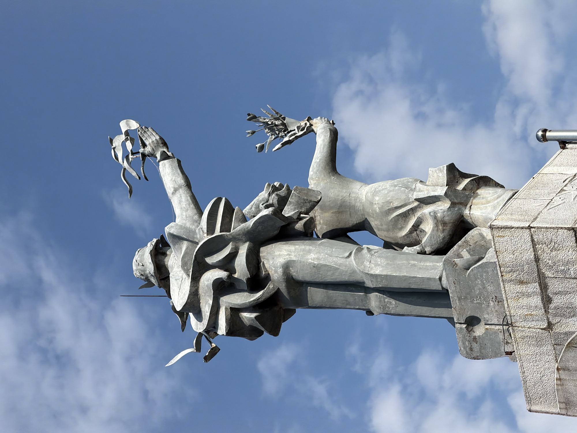 This image shows a close-up, low-angle shot of a large, abstract metal sculpture against a bright blue sky with scattered white clouds. The sculpture depicts two figures, one larger and standing, the other smaller and kneeling or crouching. The standing figure, positioned in the foreground, appears to be a man wearing a helmet and a flowing, layered garment. His right arm is raised, and in his outstretched hand, he holds a stylized bunch of three birds in flight. The smaller figure, positioned behind and below the standing figure, is also made of metal and appears to be a woman or a child. She holds a bouquet of flowers in her outstretched hand. The figures are abstract and angular, with a weathered, gray metallic finish. The sculpture is mounted on a textured stone pedestal. The overall impression is of a monument dedicated to peace or freedom.
This sculpture is the "Monument to the Revolution" or "Statue of the Revolution" located in the central square of Nha Trang, Vietnam, facing the coast. It commemorates the victory of the Vietnamese people during the revolution and their subsequent struggle for independence.