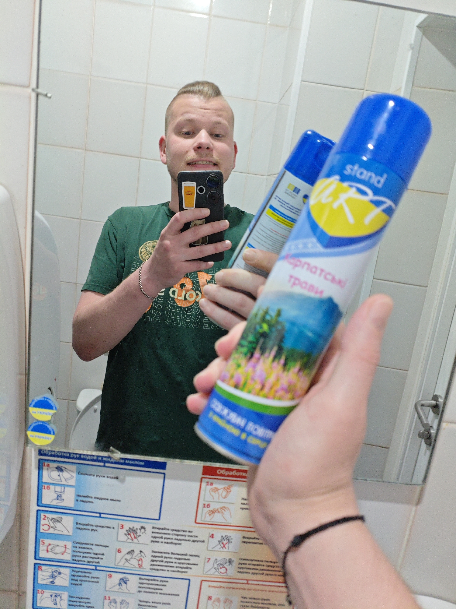The image shows a man taking a selfie in a bathroom with a mirror. He is wearing a green t-shirt and has a bracelet on his left wrist. In the mirror, the man is holding a spray can with a blue and white label that says "stand" and "Карпатські трави" (Carpathian herbs). There are two such cans reflected in the mirror. The bathroom walls are tiled in white. Below the mirror, there is a hygiene instruction poster with illustrations and text in Ukrainian, including a section on "Обробка рук водою і милом" (Hand washing with water and soap). Two circular blue and yellow stickers with the Ukrainian flag are visible on the wall to the left of the poster. The overall scene suggests a public restroom, possibly in Kyiv, Ukraine, given the Ukrainian language on the poster and stickers.