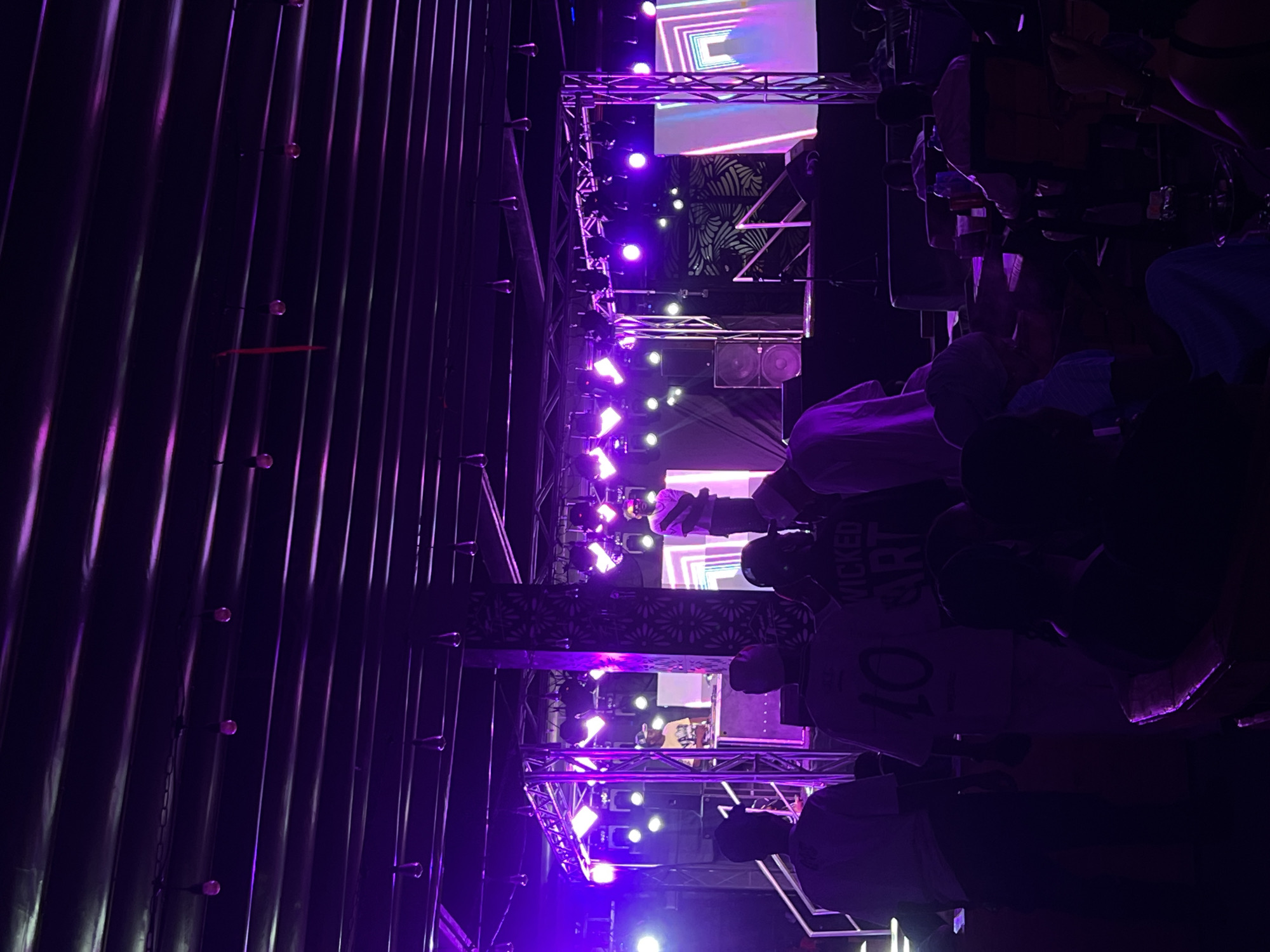 The image shows a dimly lit indoor event, possibly a concert or performance, taking place in Howayi, Nigeria. The scene is illuminated by purple stage lighting. A metal structure or frame is visible, supporting the stage setup. There are screens displaying visuals, and speakers are present. Individuals are in attendance, with some wearing shirts with visible text like "WICKED ART" and "10". People are standing and engaging with the performance. The venue appears to be enclosed by corrugated metal walls with attached light fixtures.