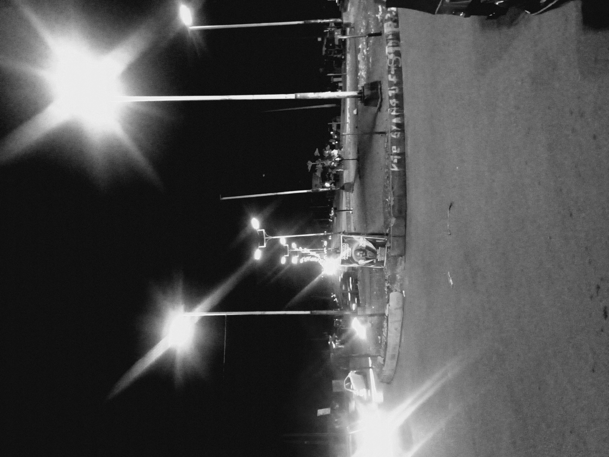 The image is a black and white photograph of a street scene at night in Bukuru, Nigeria. The focus is on a row of streetlights illuminating the area. The street is to the right with a curb marking the edge. There is writing on the side of the street. There are objects near the curb like trashcans. The streetlights are bright and create a starburst effect. There are also a few cars or vehicles visible, some of which appear to have their headlights on.