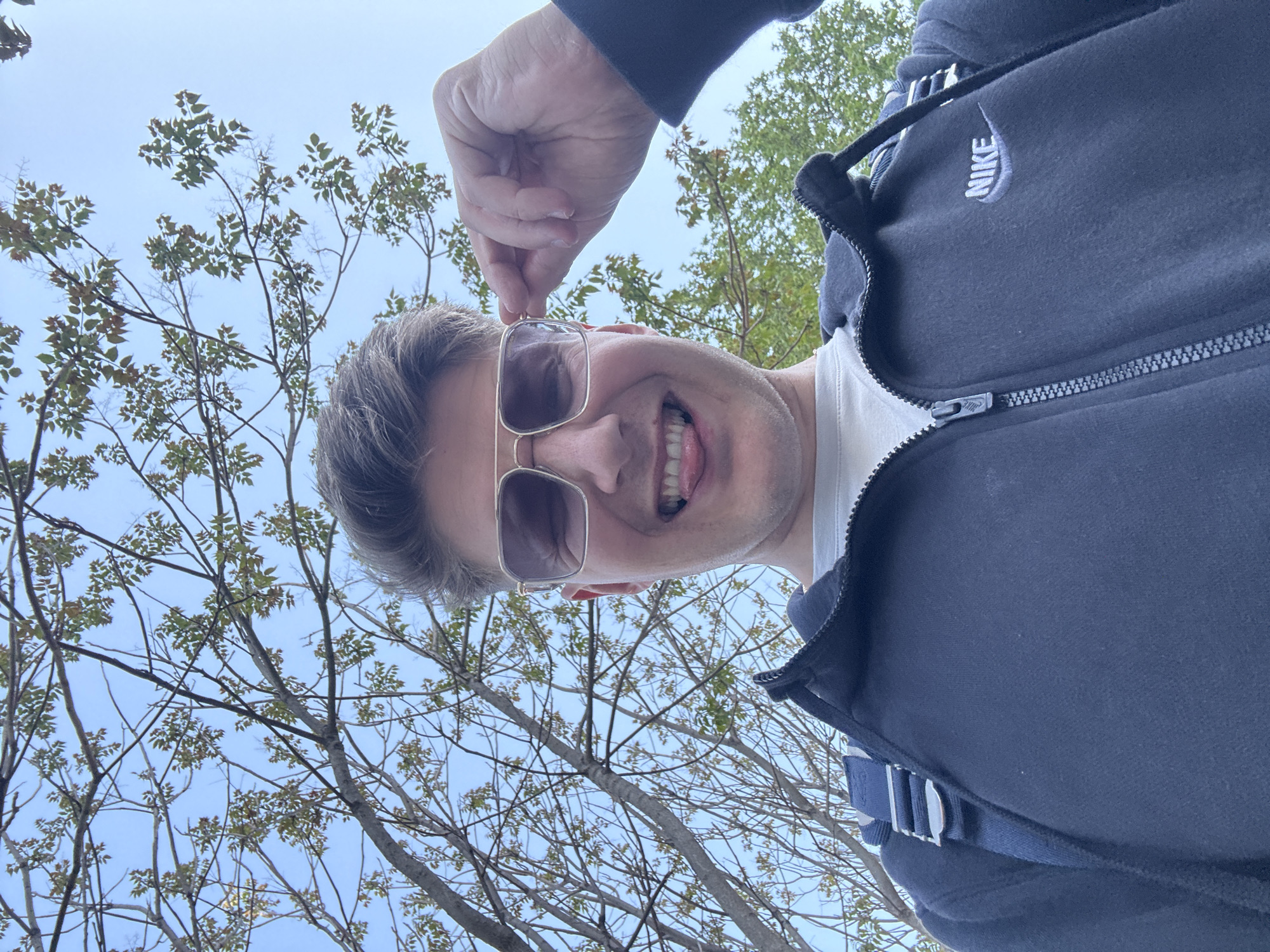 This low-angle selfie captures a man looking up towards a clear, bright blue sky in Plovdiv, Bulgaria. The man, with short, dark hair, is smiling broadly with his tongue slightly out, conveying a playful and happy mood. He is wearing aviator-style sunglasses with gold frames, which he appears to be adjusting with his left hand.
He is dressed in a dark, zip-up hooded jacket, under which a white t-shirt is visible. The prominent white "NIKE" logo is clearly visible on the right chest area of the jacket. A blue strap or bag component is also noticeable across his left shoulder/back area.
The background consists of numerous bare tree branches with sparse, light green leaves, suggesting either late winter or early spring foliage, or a specific tree species. The bright sky indicates a clear, sunny daytime setting. The overall impression is one of candid enjoyment outdoors.