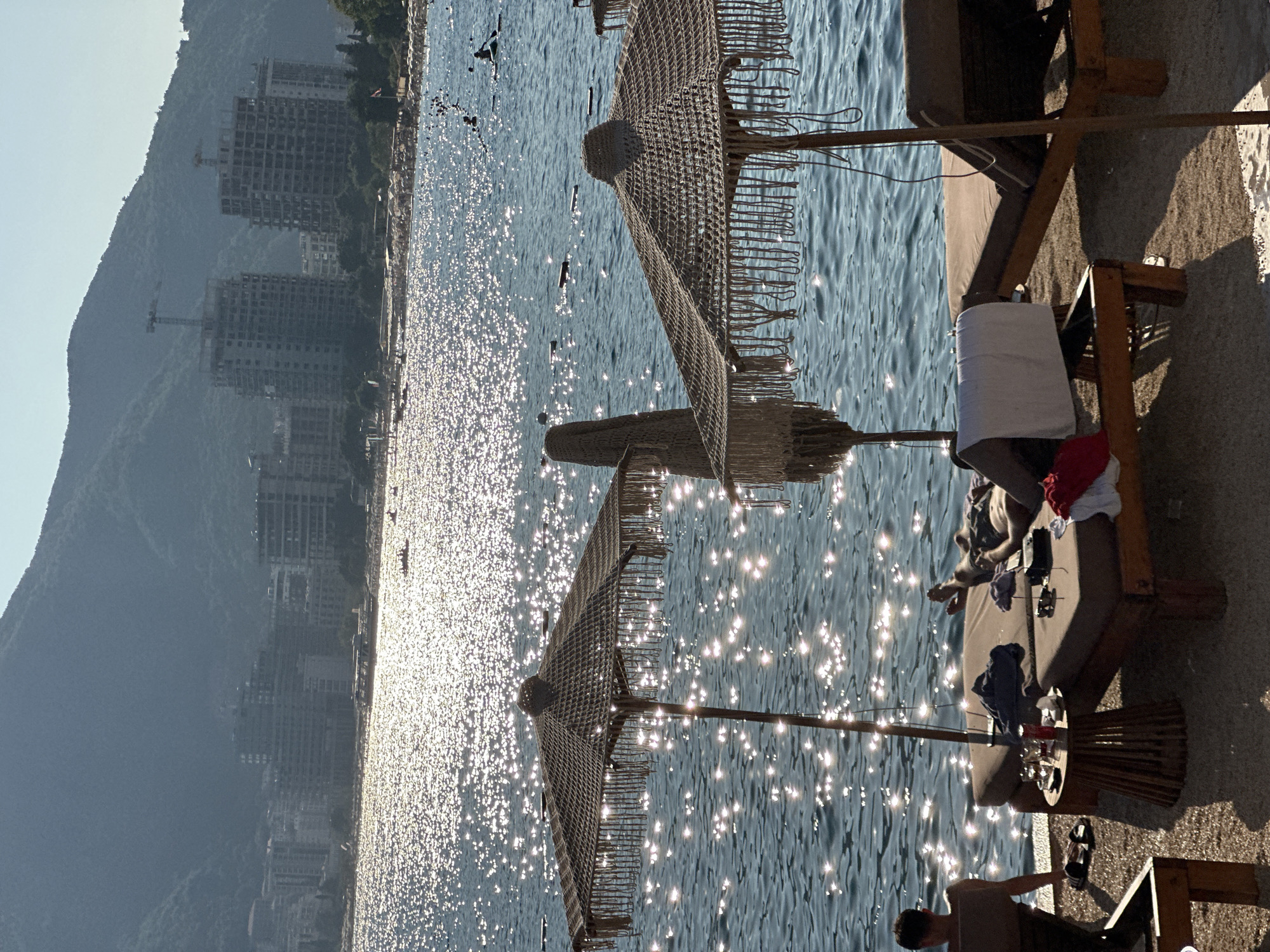 The image depicts a sun-drenched beach scene with the city of Budva, Montenegro, visible in the background. The foreground features two sun loungers, one occupied by a person reclining and the other empty. A small side table between the loungers holds drinks and a purse. Two large, thatched beach umbrellas provide shade. The sea is a sparkling blue, with numerous small boats and people enjoying water activities. In the distance, the urban landscape of Budva rises against a backdrop of lush, green mountains. The architecture suggests a mix of modern buildings and some under construction, indicative of a coastal city with ongoing development. The overall atmosphere is one of leisure and relaxation on a clear, sunny day.
