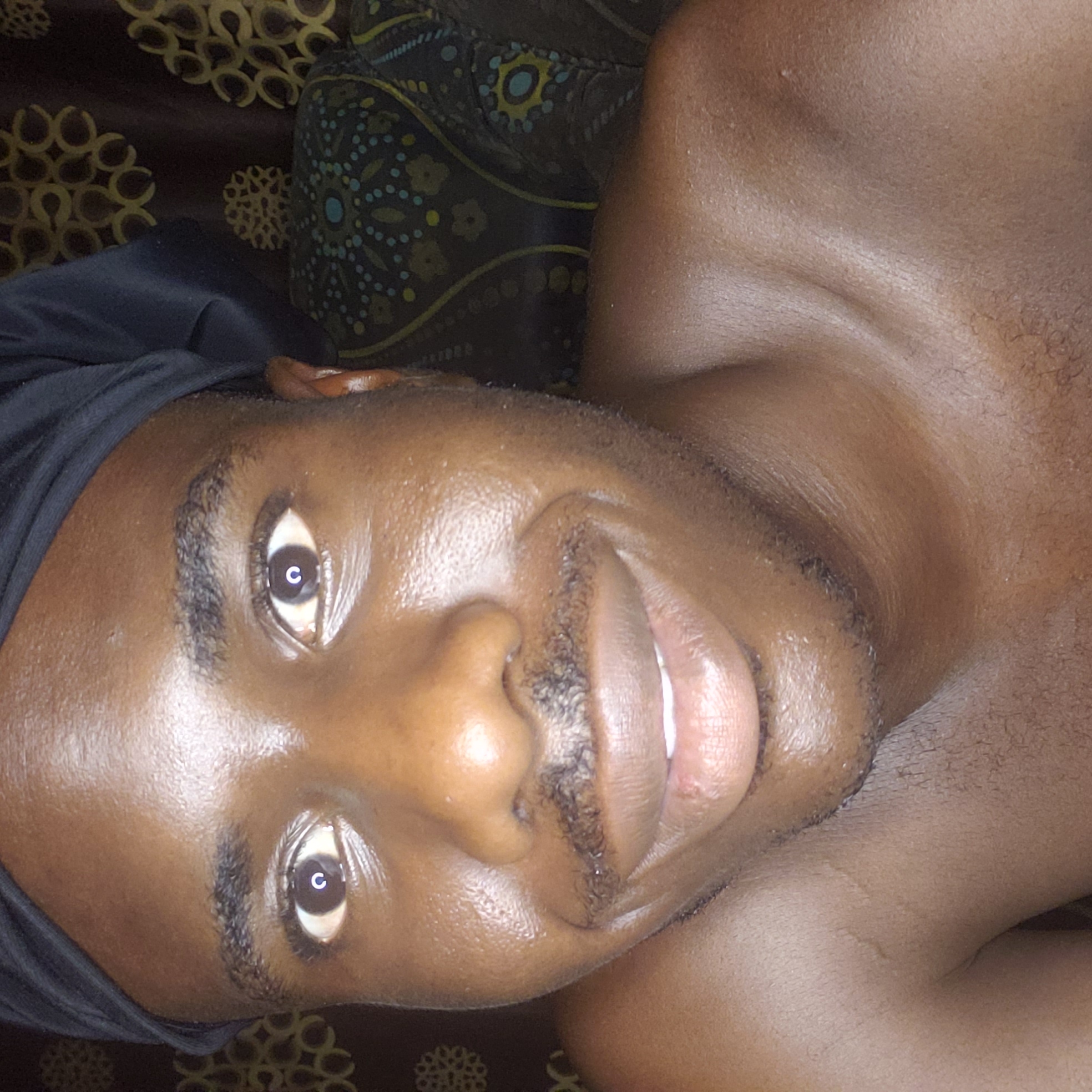 An individual with dark skin is lying on a surface covered with fabric. The person is wearing a black head covering. The individual has short, dark hair and eyebrows. The subject's eyes are open and directed toward the viewer. The lips are slightly parted, revealing teeth. The background consists of dark fabric with a repeating circular pattern. The photo was potentially taken indoors in Narayi, Nigeria.