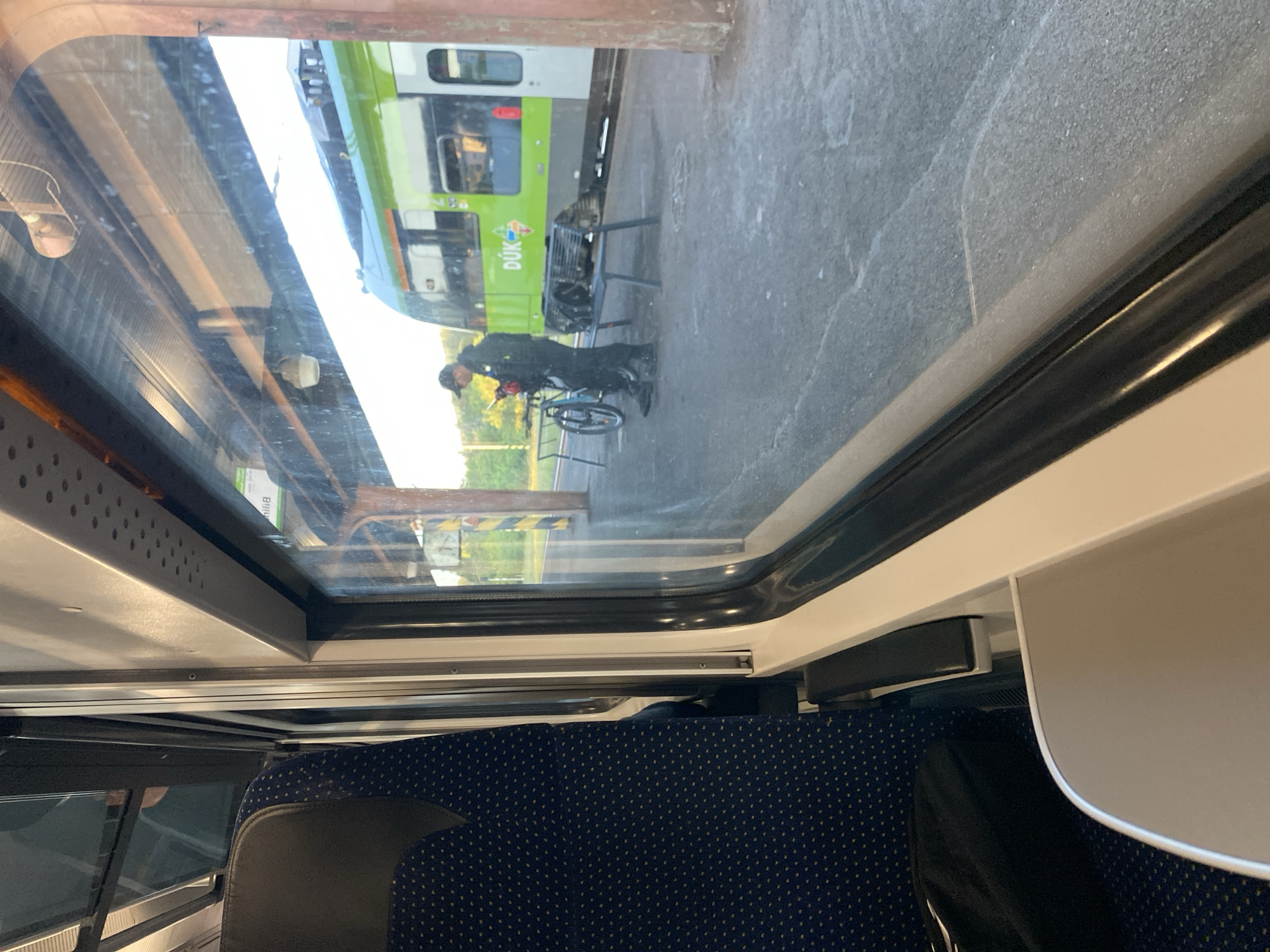 The image, taken from inside a train carriage looking out, shows a man standing on a train platform with a bicycle. He is wearing a dark jacket, dark pants, and a baseball cap. The man is holding a mobile phone in his left hand and appears to be looking at it. The bicycle is a dark color and is leaning against a bench. The train itself is green and white and is stopped at the platform. The platform is paved and there are some yellow and black striped poles in the background. The scene appears to be in Bílina, Czechia, given the train's branding includes "DÚK," which is the abbreviation for Dopravní podnik Ústeckého kraje (Transport Company of the Ústí Region), to which Bílina belongs. The time of day appears to be late afternoon or early evening, with sunlight casting long shadows.