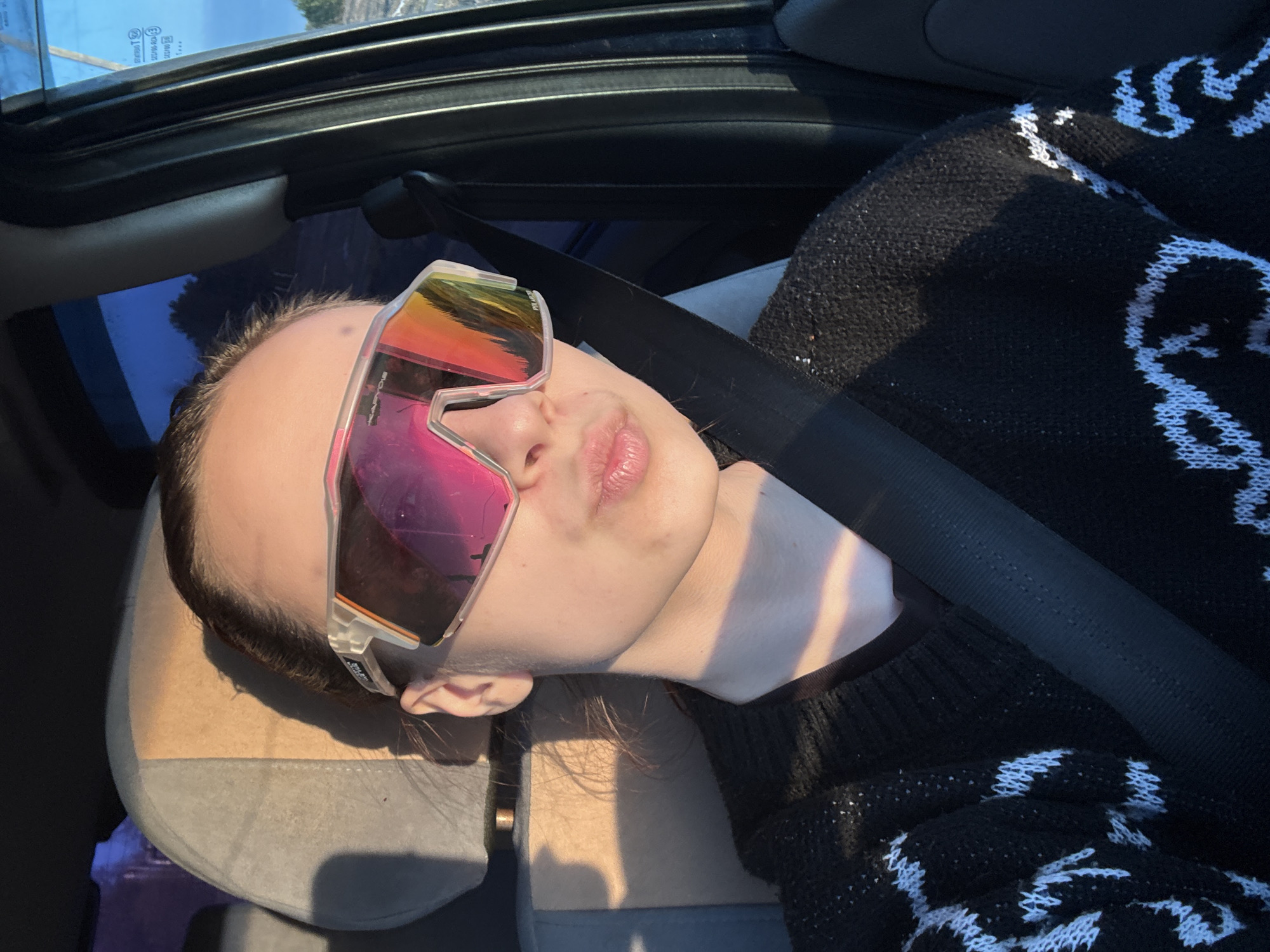 A person with fair skin is reclining in what appears to be the passenger seat of a car. They are wearing oversized, iridescent sports sunglasses with a white frame and a black and white patterned sweater. A black seatbelt is fastened across their torso. The headrest is gray and the interior of the car is visible, including the sunroof. The location of Прикамский, Russia cannot be determined from the image.