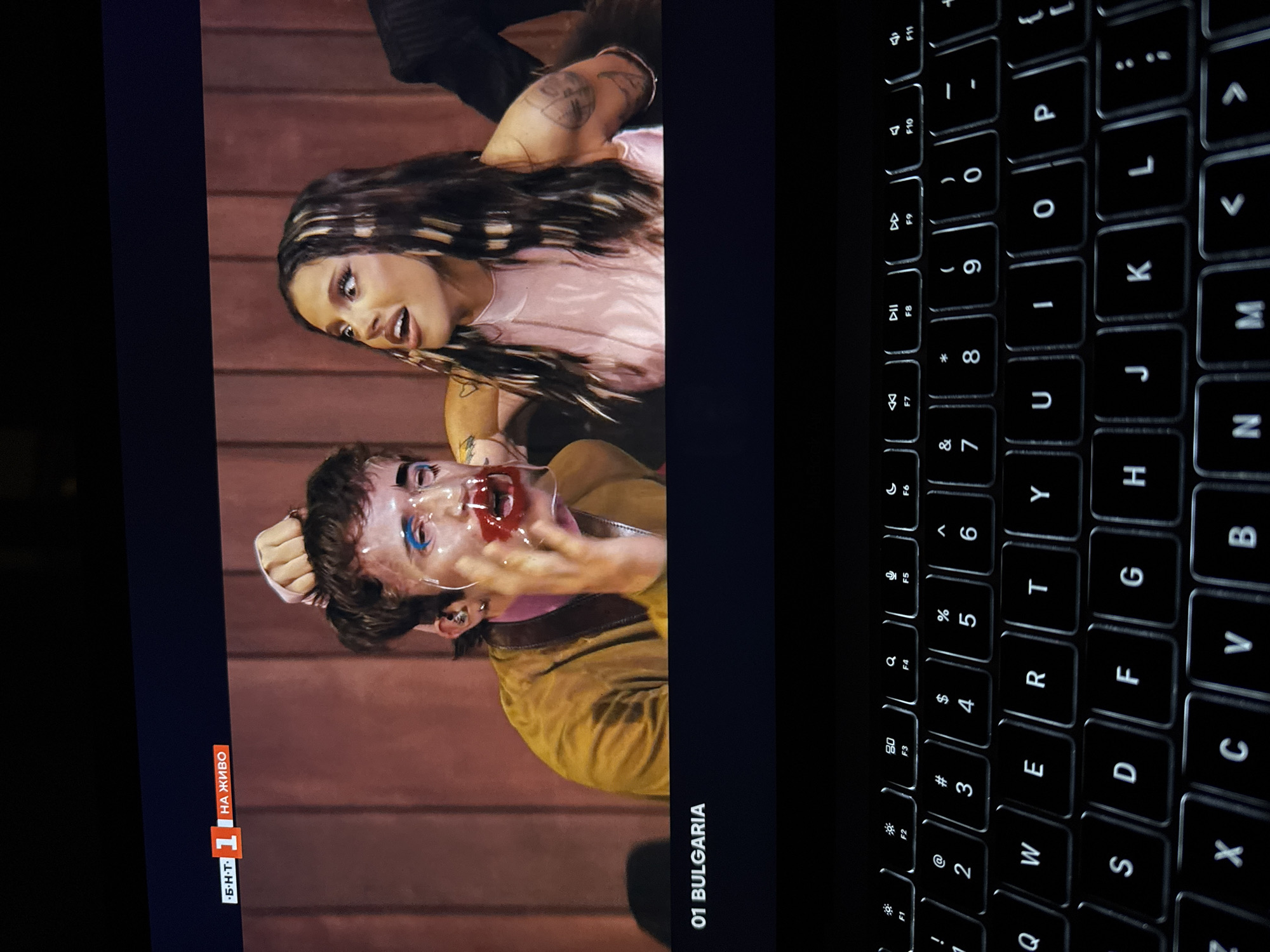 The image shows a scene being viewed on a laptop, with the keyboard visible on the right. The screen displays a TV show, possibly a competition or performance related to Bulgaria, as indicated by the text "01 BULGARIA" and "НА ЖИВО" (translated as "LIVE"). The image depicts two individuals: one with a mask or face makeup that seems to be smeared with red, possibly blood, and the other, a woman with long dark hair with blonde streaks and a tattoo on her arm. They are positioned against what looks like a wood-paneled background. The specific location is not identifiable in the photo and there is no indication of it being in the city of Milan, Italy.
