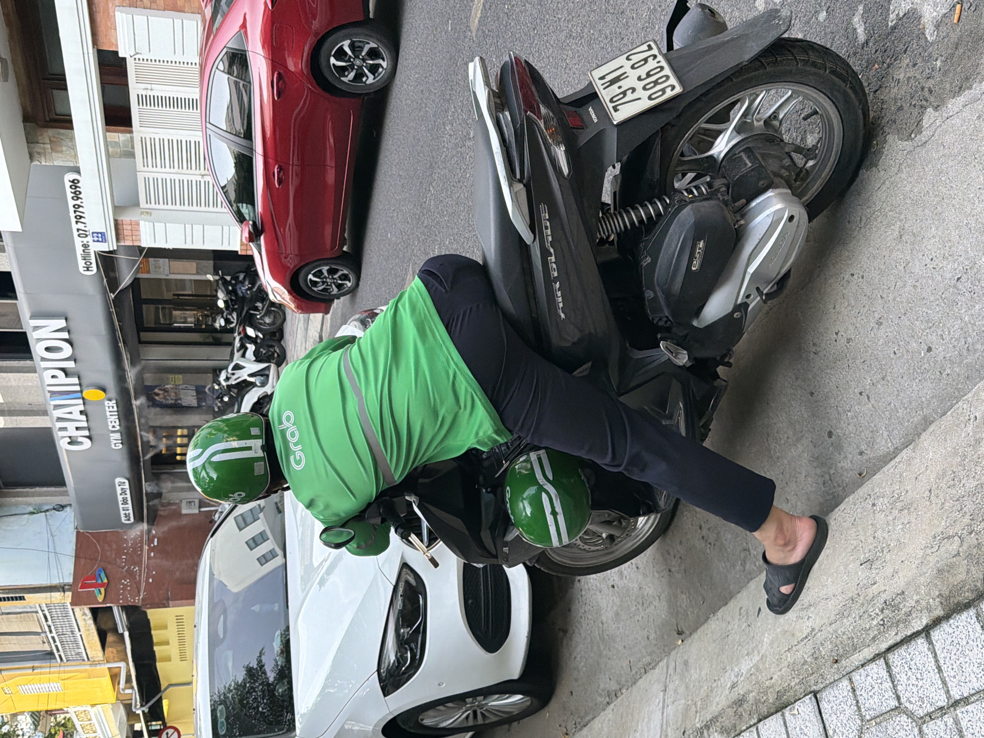 In Nha Trang, Vietnam, a Grab delivery driver, wearing a green helmet and uniform, is positioned on a black motorcycle. The driver's left leg is extended, resting on a concrete curb, and they are leaning forward on the handlebars. A white car is parked to the left of the motorcycle, and a red sedan is visible in the background. Behind the driver, a sign reads "CHAMPION GYM CENTER" with a hotline number. The scene appears to be a street in a commercial area.