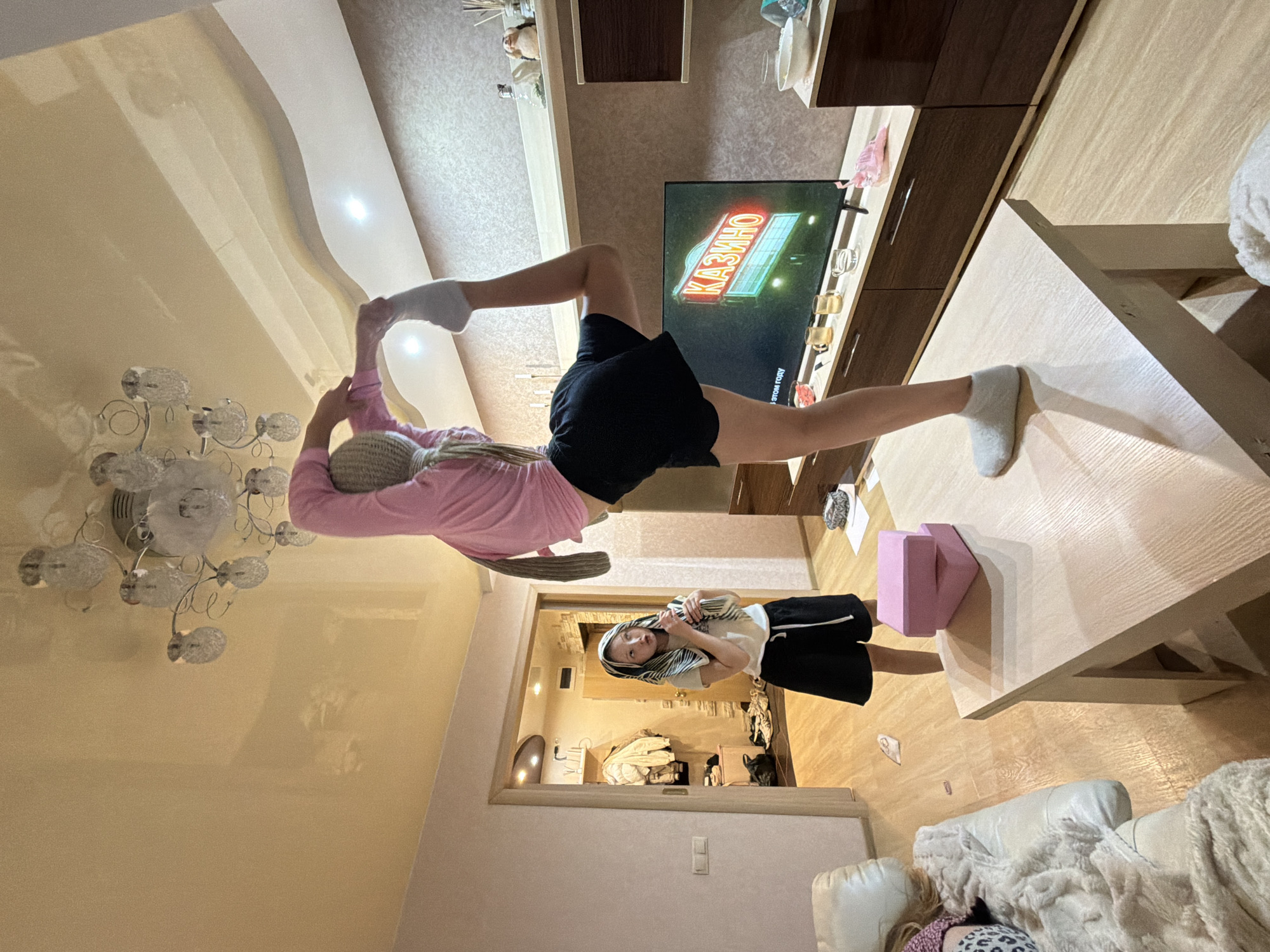 A girl in a pink long-sleeved shirt, black shorts, and a beige hat is performing a yoga pose in a living room. She is standing on a coffee table with pink blocks and balancing with one leg extended towards a television screen displaying the word "Казино" (Casino) in Russian. Another girl with short hair and a white top with black shorts, wearing a striped scarf, is looking at the camera. The room contains furniture, including a TV stand, a sofa, and a chandelier. The setting appears to be a domestic interior, likely in an apartment in the city of Chaikovsky, Russia.