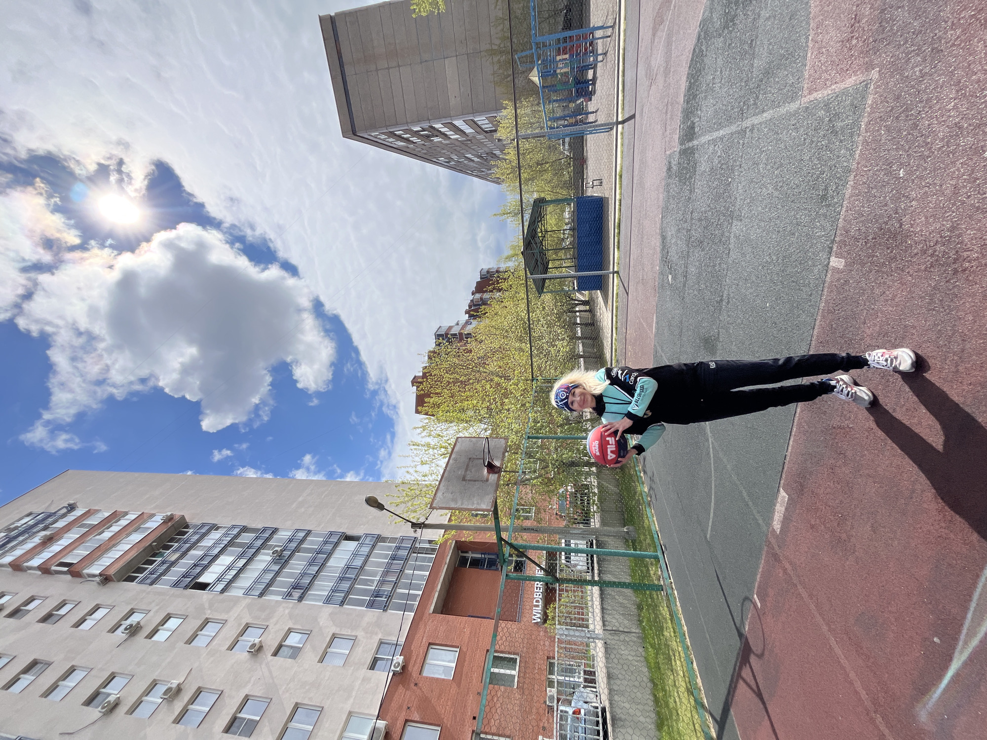 A woman stands on a basketball court in Krasnoyarsk, Russia, holding a basketball. The court is bordered by buildings, one of which has air conditioning units on the exterior walls. A basketball hoop is visible. The sky is blue with clouds and a bright sun.