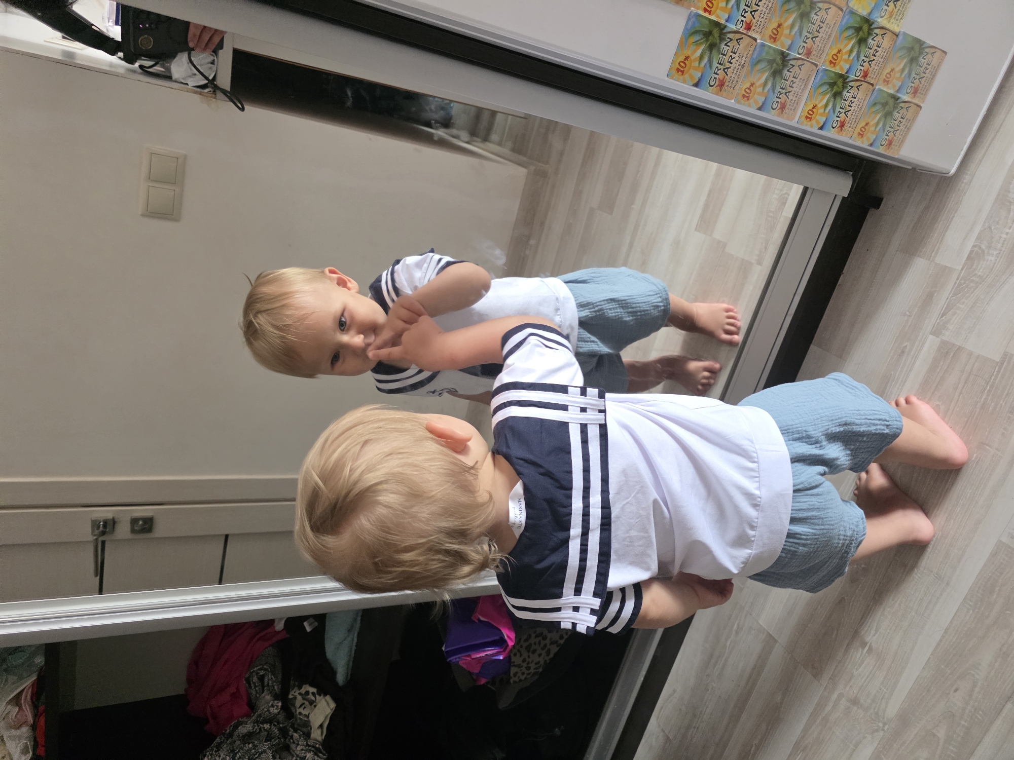 A toddler with blonde hair and blue eyes, wearing a white sailor-style shirt with blue trim and light blue shorts, is looking into a full-length mirror. The child is touching their finger to their nose. The toddler is standing on a light wood-grain floor. To the left of the toddler is a closet with clothes visible inside. To the right of the toddler is a white refrigerator with several small square magnets attached to its surface. The magnets appear to be advertisements for "GREEN AREA" with palm trees and a 10% discount indicated. The scene is indoors, likely in a residential setting. The city of Sochi, Russia, is not depicted in the image.