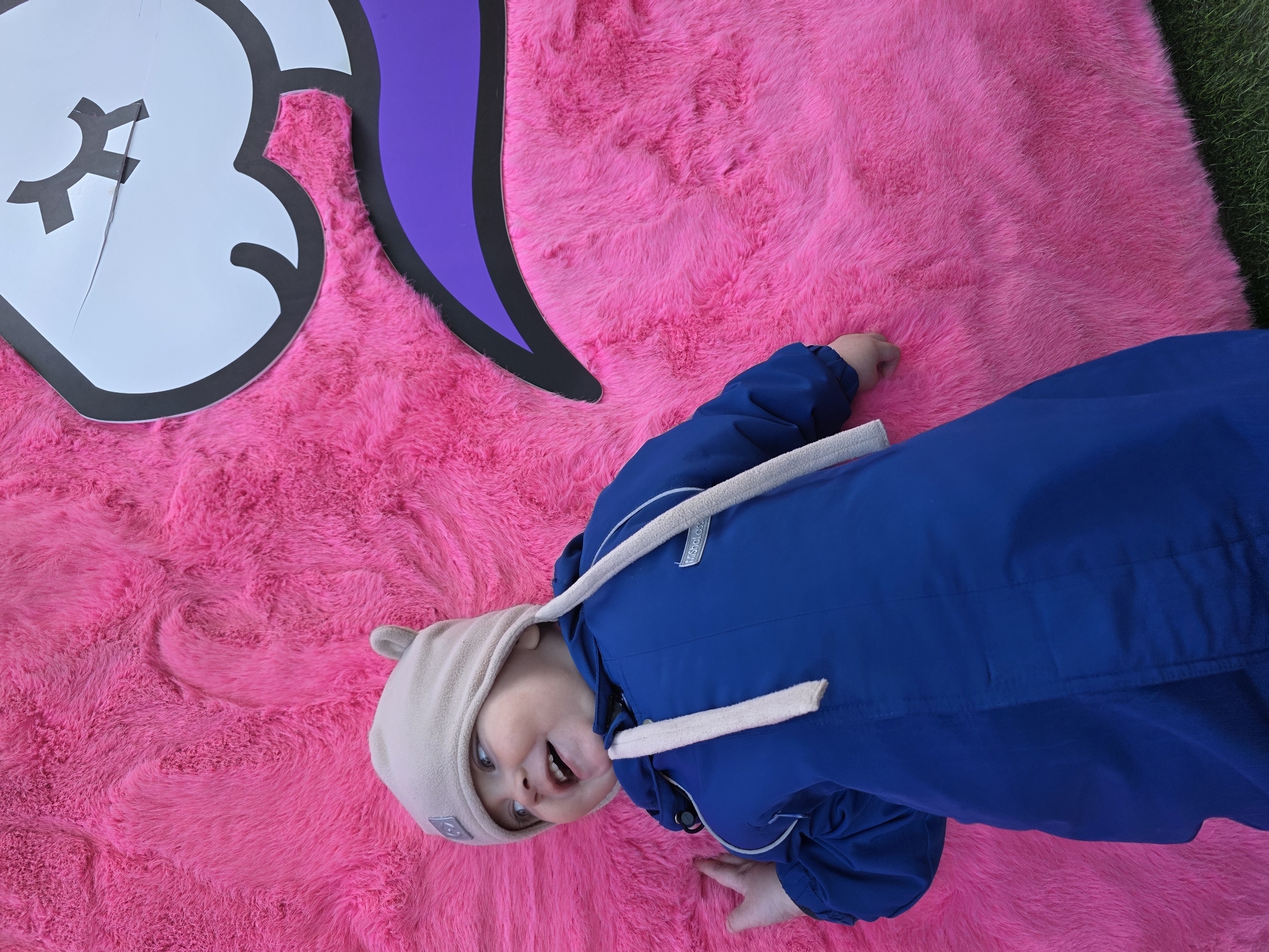 A young child is depicted lying on their back on a large, bright pink faux fur surface in Votkinsk, Russia. The child, wearing a royal blue long-sleeved jacket with a "MiShaLoss" label on the right sleeve and a light beige beanie featuring small ear-like protrusions and dangling ties, looks upward and to the left with an open-mouthed smile, revealing teeth. A light grey square patch with an emblem is visible on the beanie. In the upper right quadrant, a portion of a large white cartoon character's face, outlined in black, is visible. It features a stylized closed eye represented by a cross shape and a curved purple element, integrated with the pink fur, which appears shaggy. A strip of green grass is visible along the bottom right edge of the frame, beneath the pink surface.