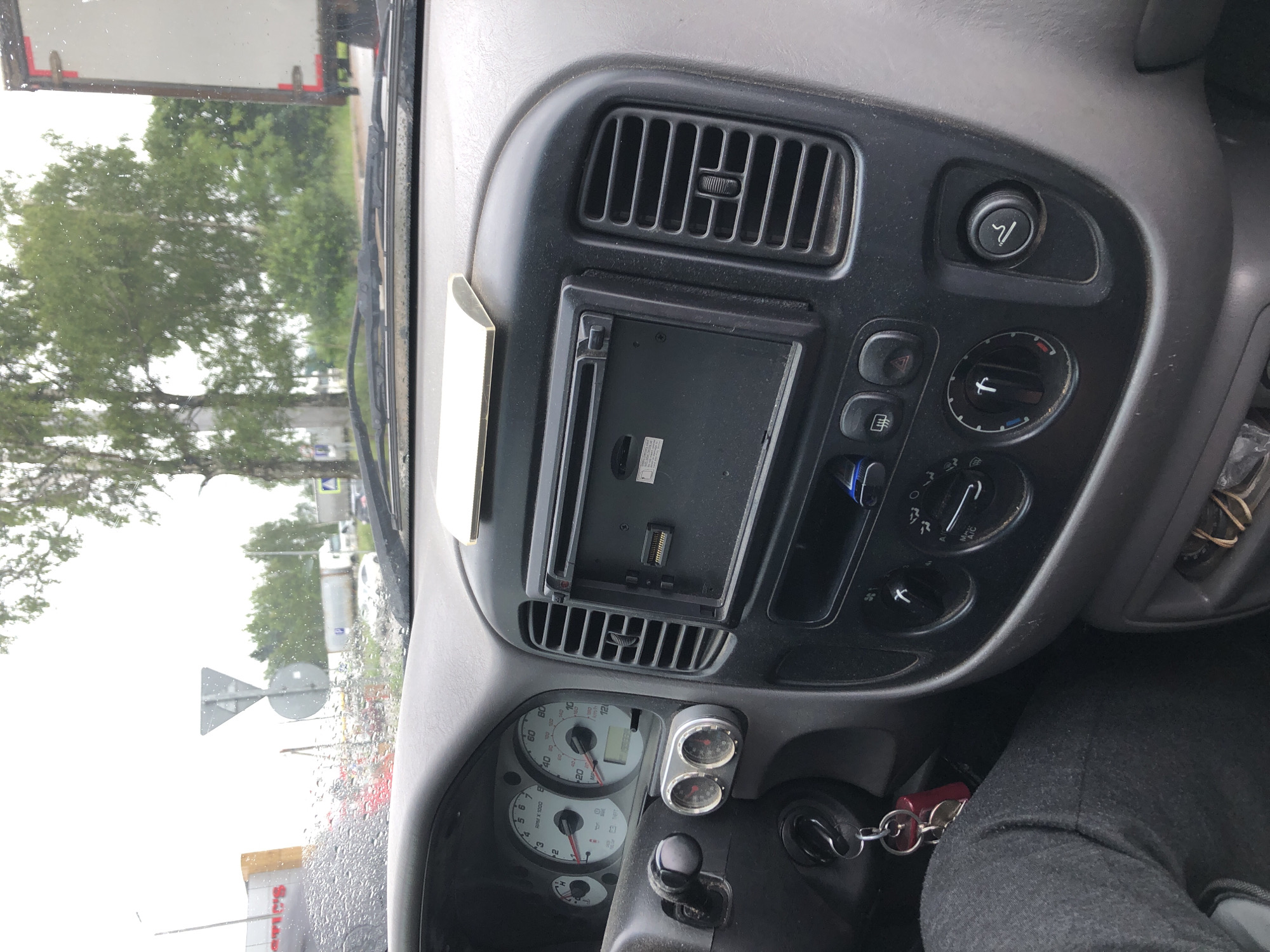 The image shows the interior of a car's dashboard and windshield, with a view of the outdoors through the rain-streaked glass. The dashboard features a radio or navigation unit, air vents, climate controls, a steering wheel with a key in the ignition, and gauges for RPM and speed. Outside, trees, a road, and a truck are visible. The scene appears to be in an outdoor urban or semi-urban environment, possibly Safonovo, Russia, given the context. The weather is overcast and rainy, as indicated by the water droplets on the windshield and the general lighting. There are no people clearly visible within the car.