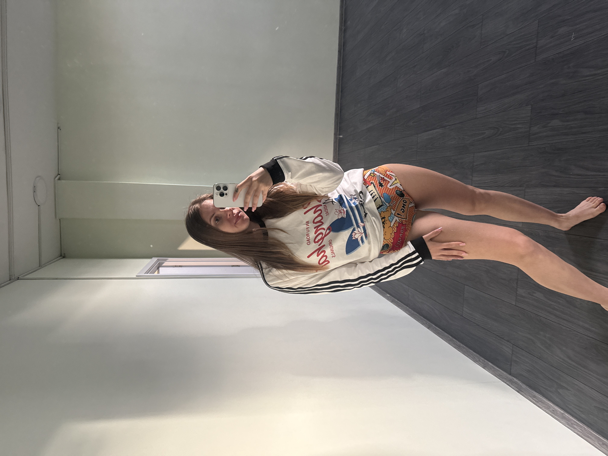 A young woman is taking a selfie in what appears to be an apartment or studio. The background consists of light-colored walls and dark grey wood-look flooring. There is a window in the background. The woman is wearing a white sweatshirt with black stripes on the sleeves and patterned shorts. She is holding a smartphone in her right hand to capture the photo. The location appears to be indoors and does not offer any clues as to the city of Batumi.