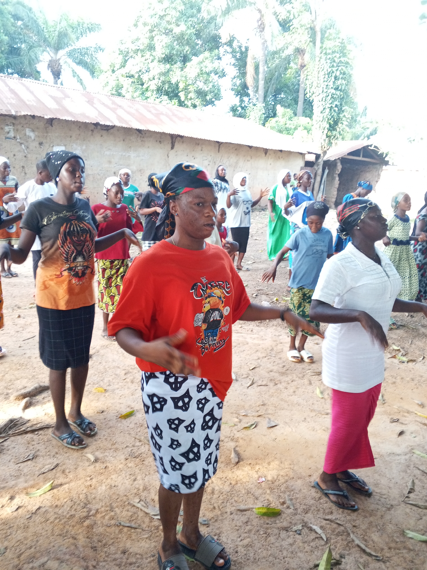 A group of women are gathered in an open, sandy area in Akwanga, Nigeria, possibly a village setting. They are predominantly dressed in casual clothing, including t-shirts and skirts, with some wearing headscarves or wraps. They appear to be engaged in some sort of synchronized movement or exercise, with their arms extended to the sides. The environment includes simple buildings constructed with corrugated metal roofing and trees in the background. The scene suggests an outdoor activity or gathering within a rural or semi-rural area.