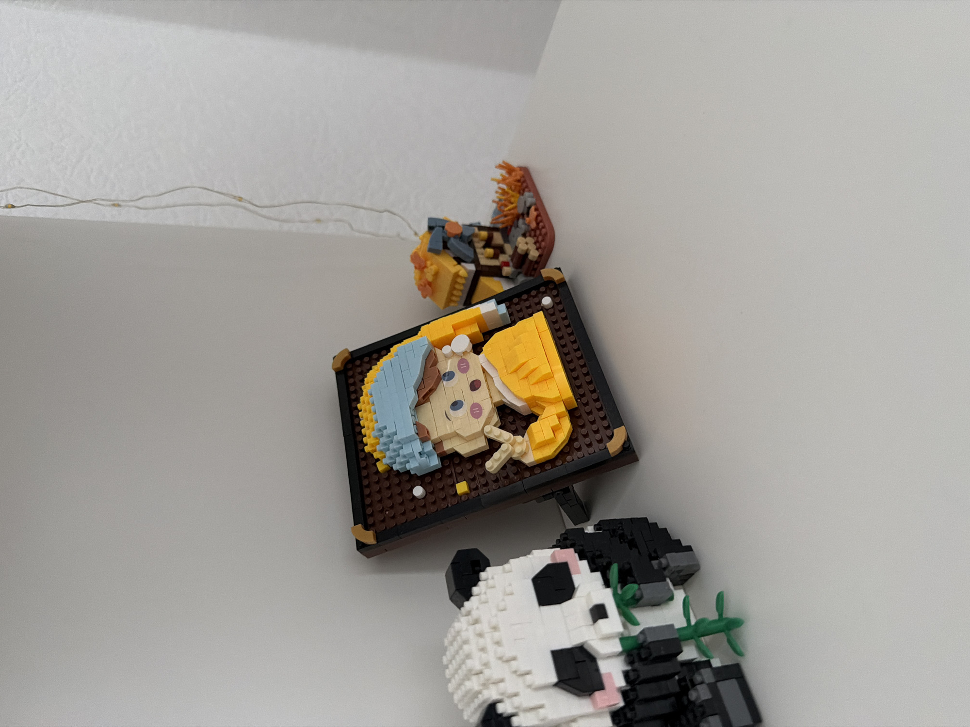 On a white shelf in Kyiv, Ukraine, a collection of miniature brick-built figures is displayed. To the left, a panda figure made of white and black bricks is depicted munching on green bamboo stalks. Next to it, a framed portrait, also constructed from bricks, features a young person with a blue headscarf and yellow clothing, holding up two fingers in a peace sign. The portrait resembles Vermeer's "Girl with a Pearl Earring." To the right of the portrait, a small brick-built structure, appearing to be a house with a yellow roof, is surrounded by orange and red brick elements suggesting flames or autumn leaves. A strand of delicate fairy lights with small yellow bulbs hangs vertically against a pale wall in the background.
