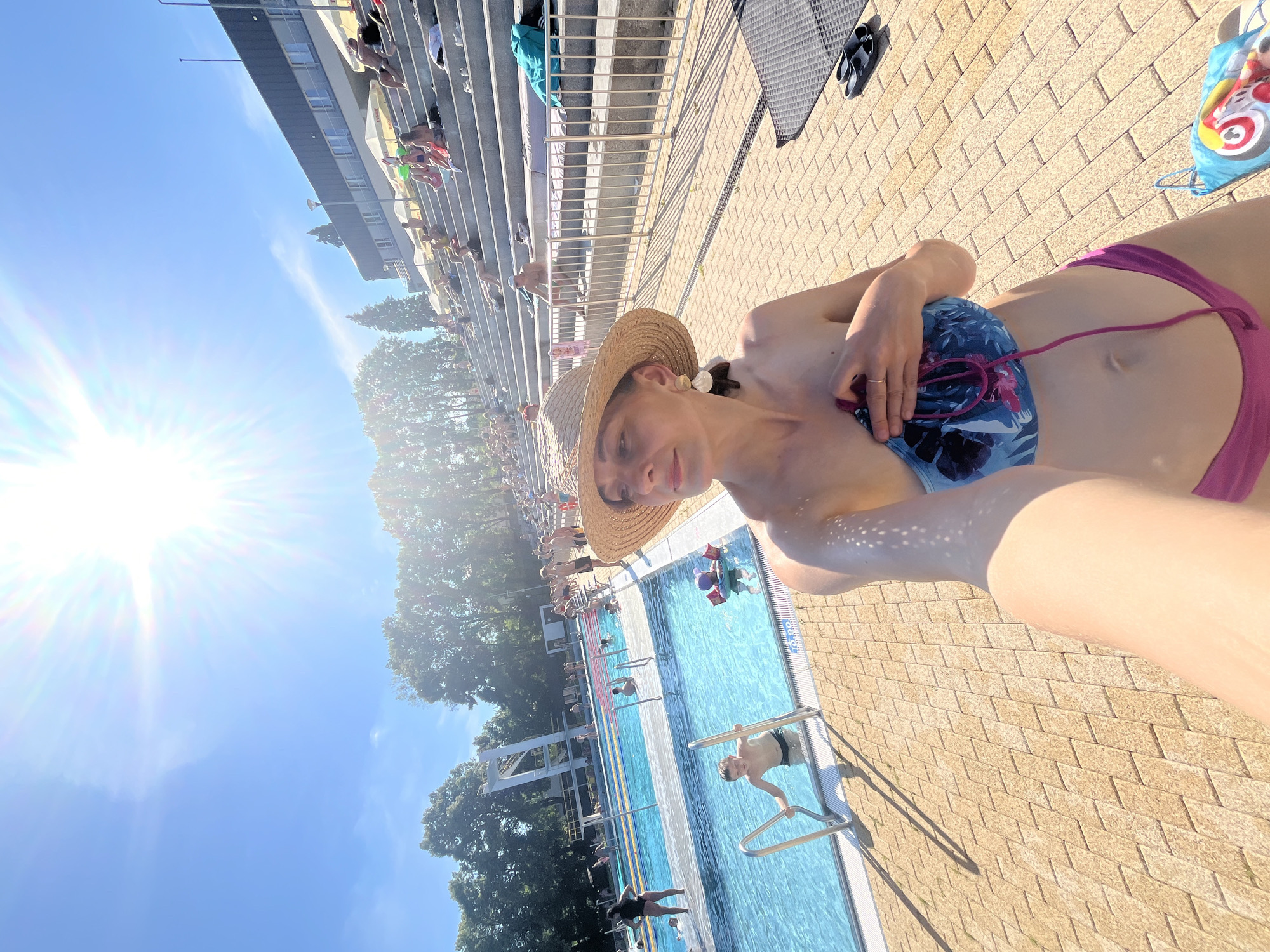 The image depicts a sunny day at an outdoor swimming pool complex. The main subject is a person wearing a straw hat and a bikini, taking a selfie. Their arm and torso are prominently featured in the foreground. In the background, there is a swimming pool with several people swimming and lounging. Some are visible on the steps leading up to the pool. Trees and a building are also visible in the distance, along with the bright sun in the clear blue sky. The ground surrounding the pool is paved with light-colored bricks. Shadows indicate a high sun, suggesting midday. The overall mood appears relaxed and summery. There is no visible text in the image.