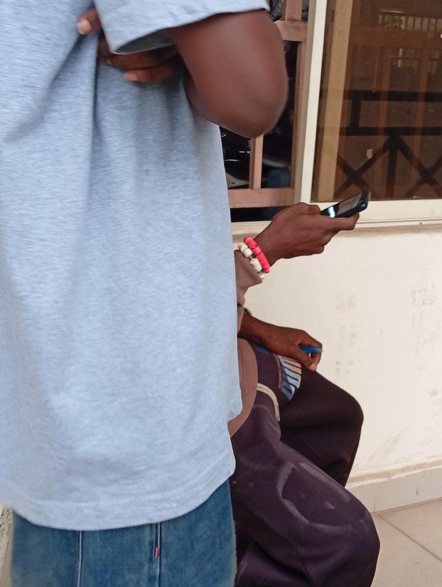 The image shows a close-up view of two individuals in Ukukwa, Nigeria. The person in the foreground is wearing a light gray t-shirt and blue jeans. The person in the background, who is seated, is wearing a dark brown tracksuit. The seated person is holding a smartphone in their right hand and a blue pen in their left hand, with two bracelets on their right wrist. They appear to be looking at the phone. The background consists of a wall and a window with a wooden lattice. The lighting suggests it is daytime.